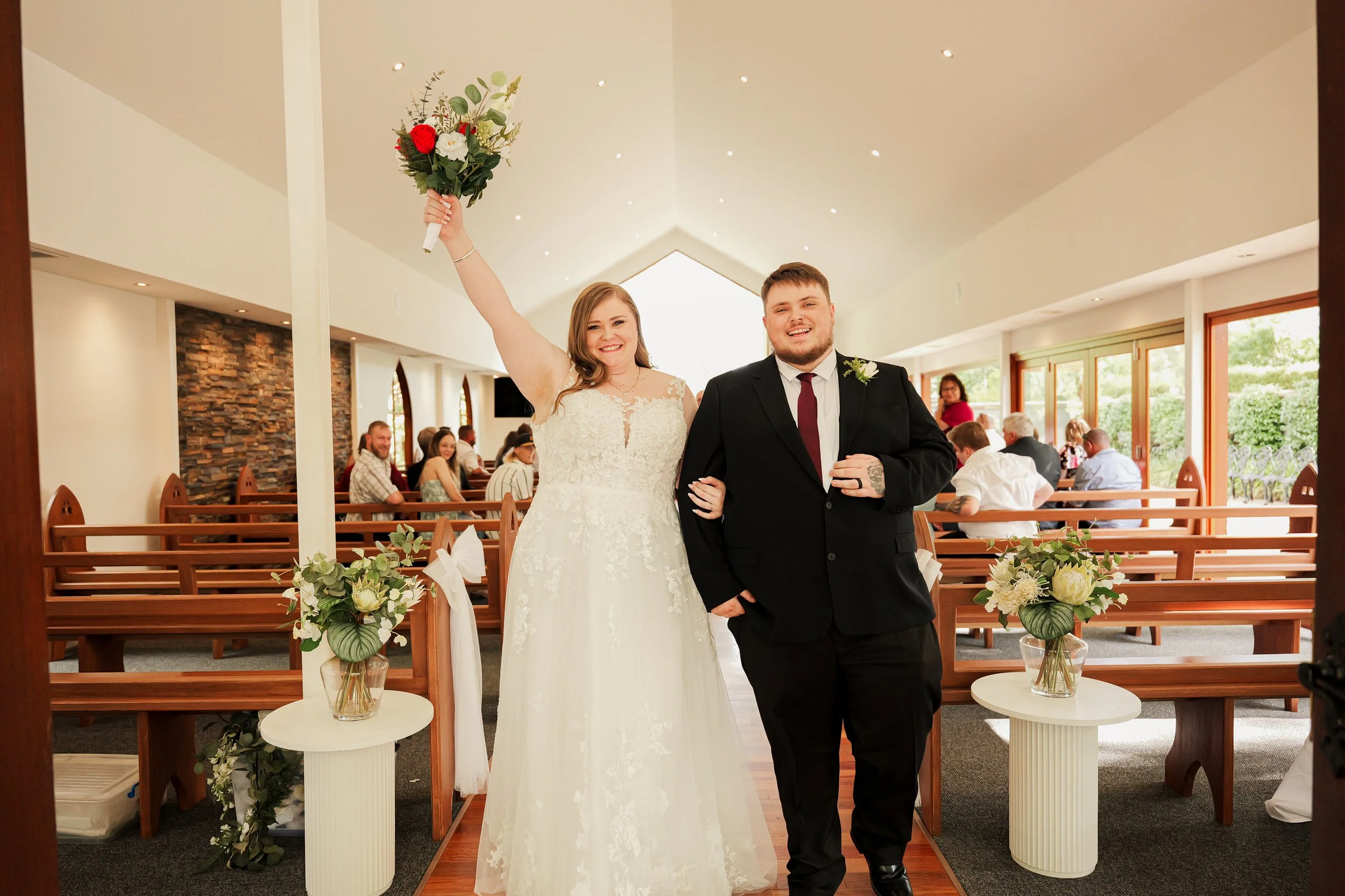A bride and groom smiling inside a church or wedding venue. The bride is holding a bouquet of flowers in her raised hand, and the groom is standing next to her with a boutonniere. People are seated in the background.