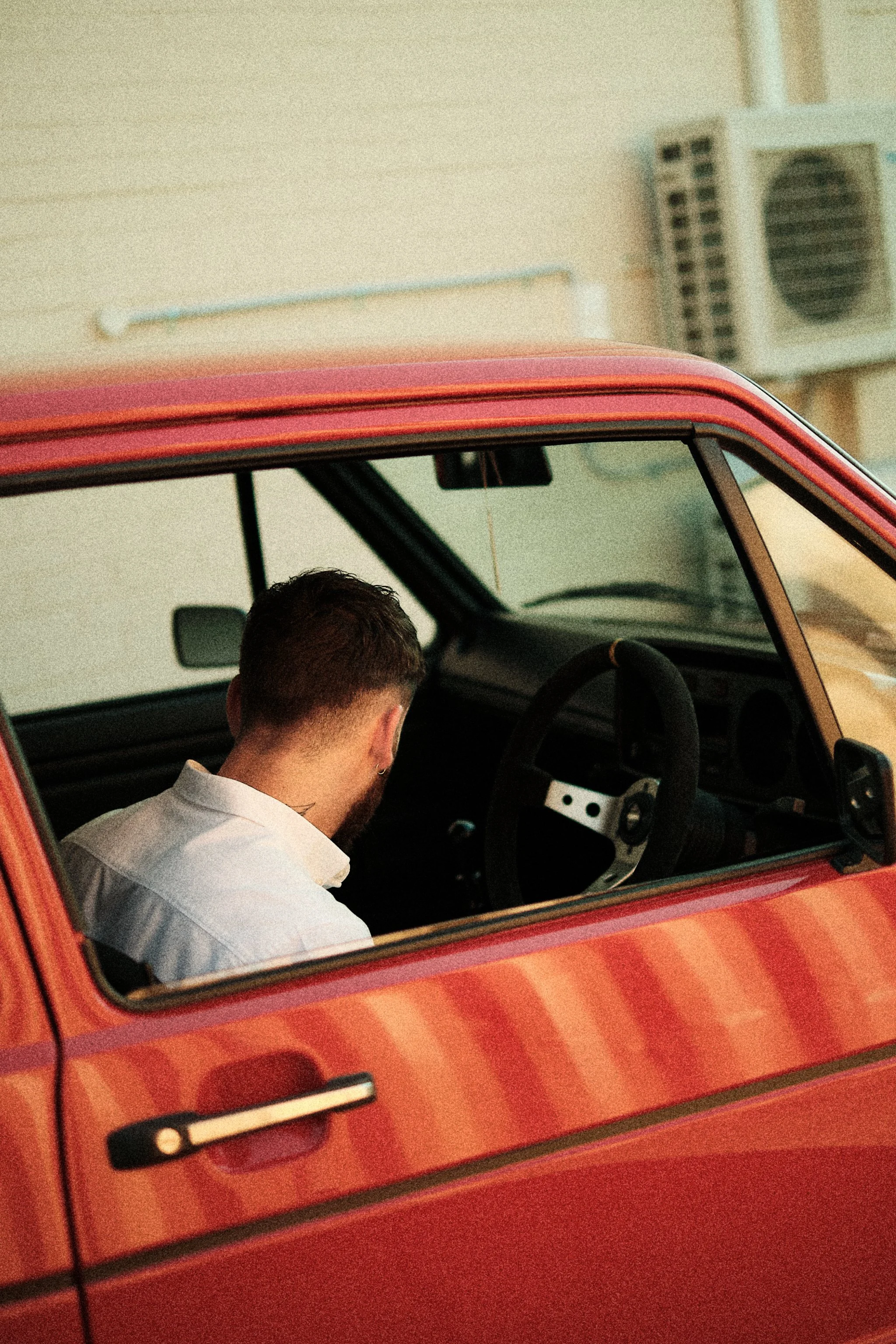 A person with short hair and a white shirt sitting inside a red vintage car, viewed through the open window. The background shows a beige wall with an air conditioning unit.