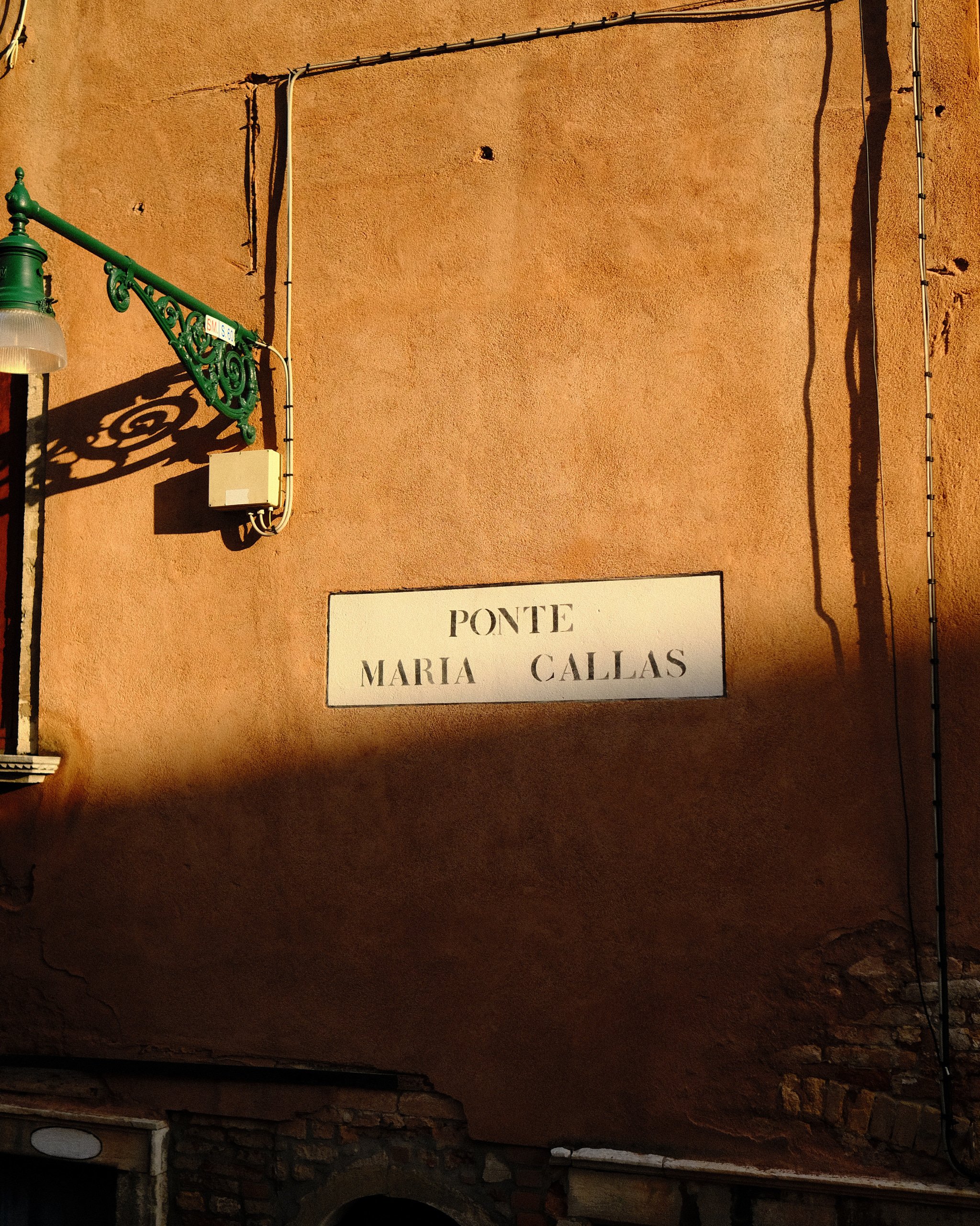 A street sign on a brick wall reading "PONTE MARIA CALLAS" with a decorative green street lamp casting a shadow, electrical wiring, and a portion of an archway at the bottom.