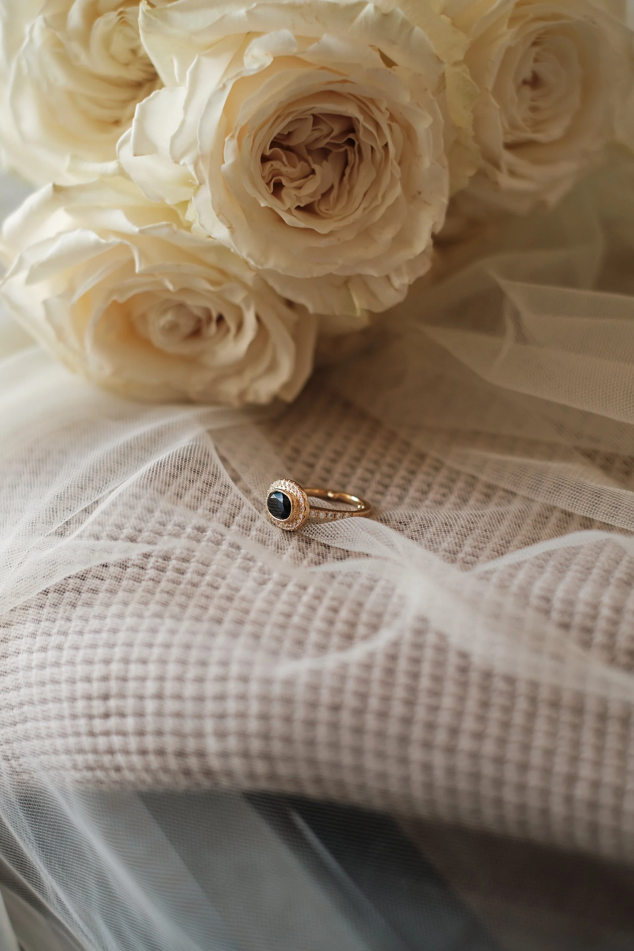 Close-up of a bouquet of cream-colored roses with a gold ring featuring a black gemstone resting on a sheer white fabric.
