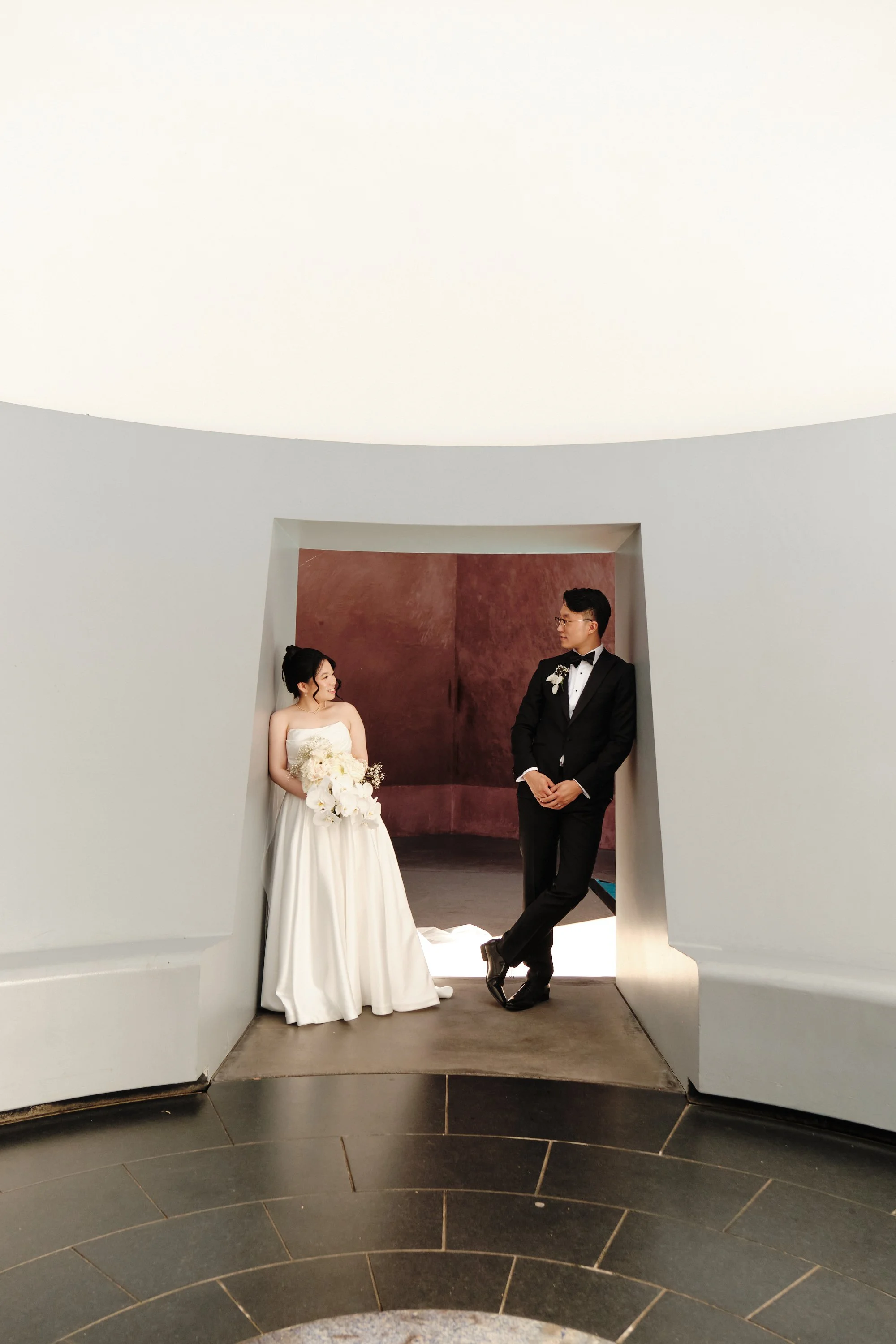 A bride and groom standing inside a modern, minimalist building with curved white walls and a dark red background. The bride is wearing a white strapless wedding gown and holding a bouquet, while the groom is dressed in a black tuxedo with a bow tie 