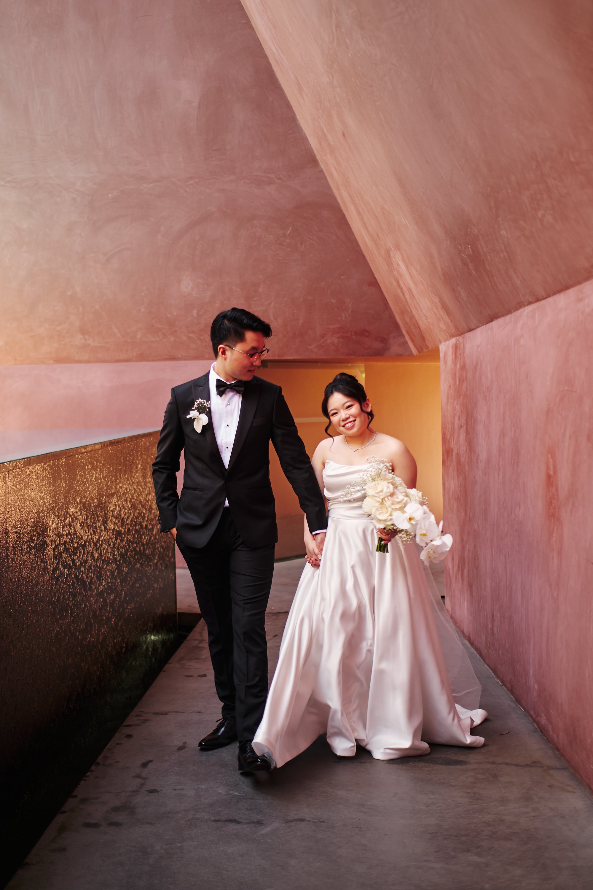 A bride and groom walking together in a hallway with pink and gold walls. The groom is in a black tuxedo with a white shirt, bow tie, and boutonniere. The bride is in a strapless white gown holding a bouquet of white flowers, smiling at the camera.
