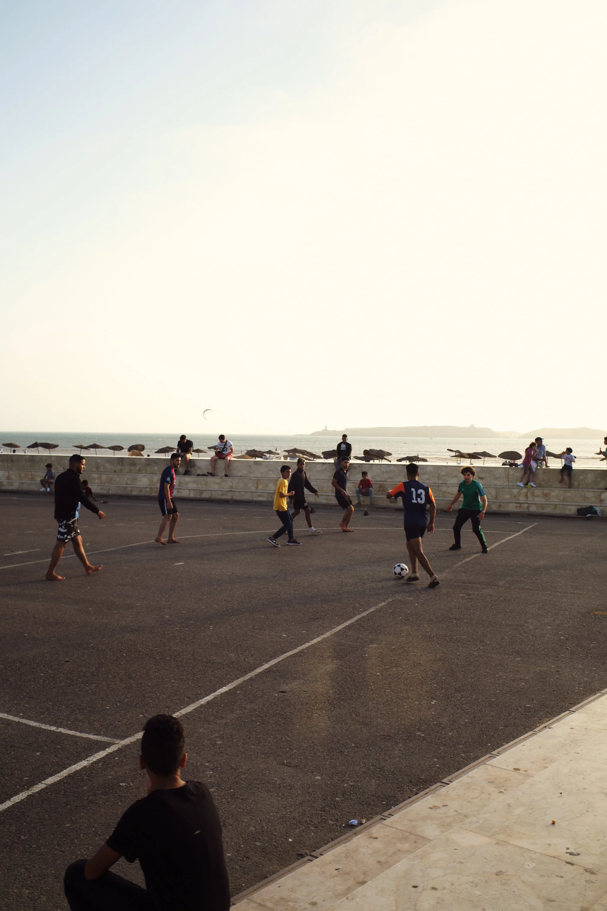 Children and adults playing soccer on an outdoor court near the beach with umbrellas, a stone wall, and the ocean in the background during the evening.