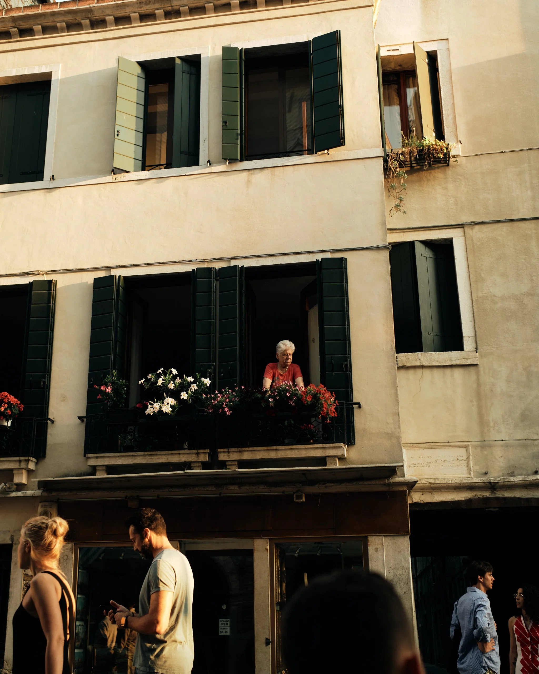 City street scene with a building featuring multiple open green shutters and a woman on a balcony with flowers, and several people walking and talking on the sidewalk.
