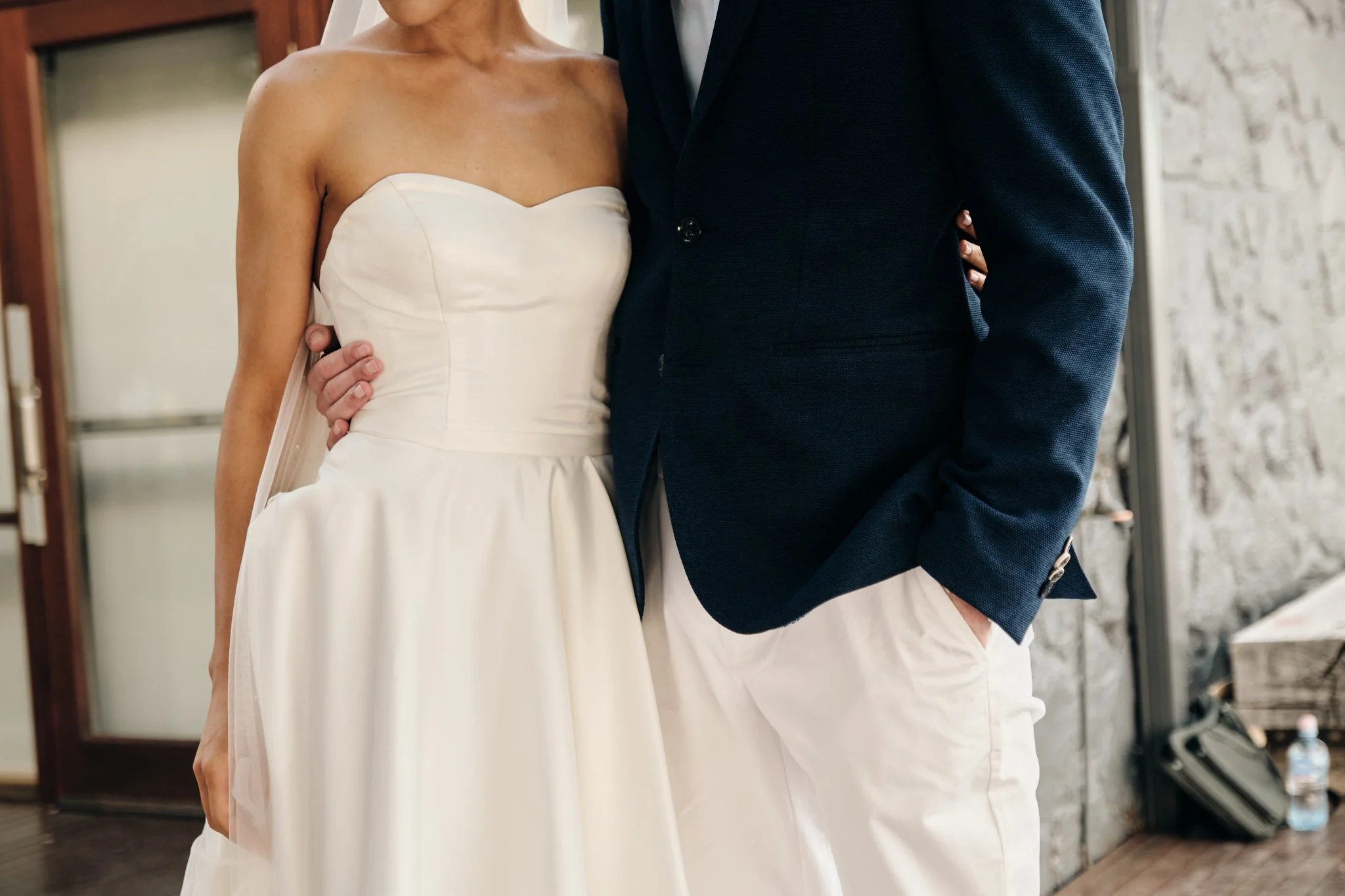 Close-up of a bride and groom during a wedding ceremony, with the bride in a strapless white wedding gown and the groom in a dark textured suit, standing indoors against a stone wall background.