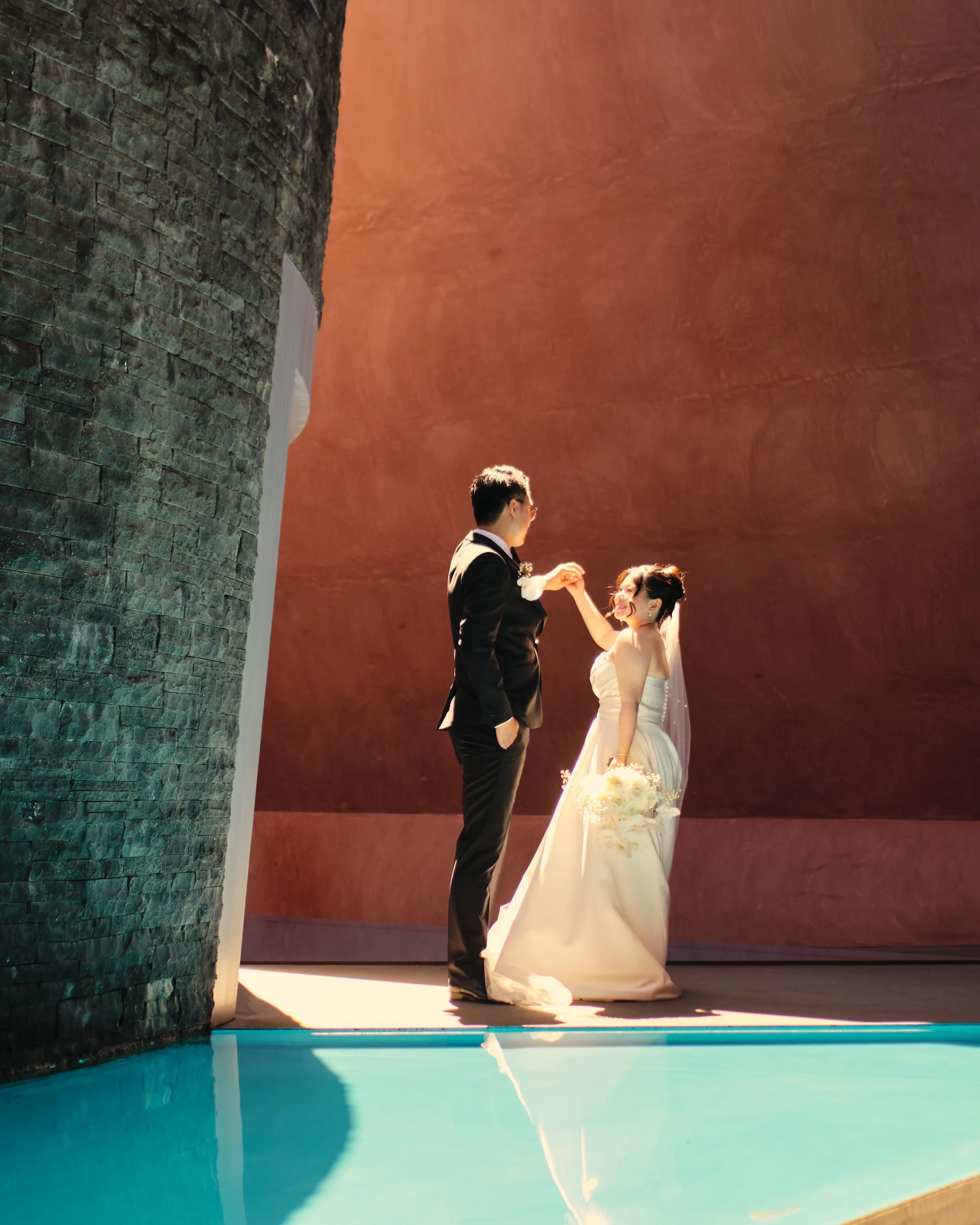 A couple dressed in wedding attire holding hands and smiling during a wedding ceremony in front of a large stone wall with a waterfall feature and a pool of water reflecting the scene.