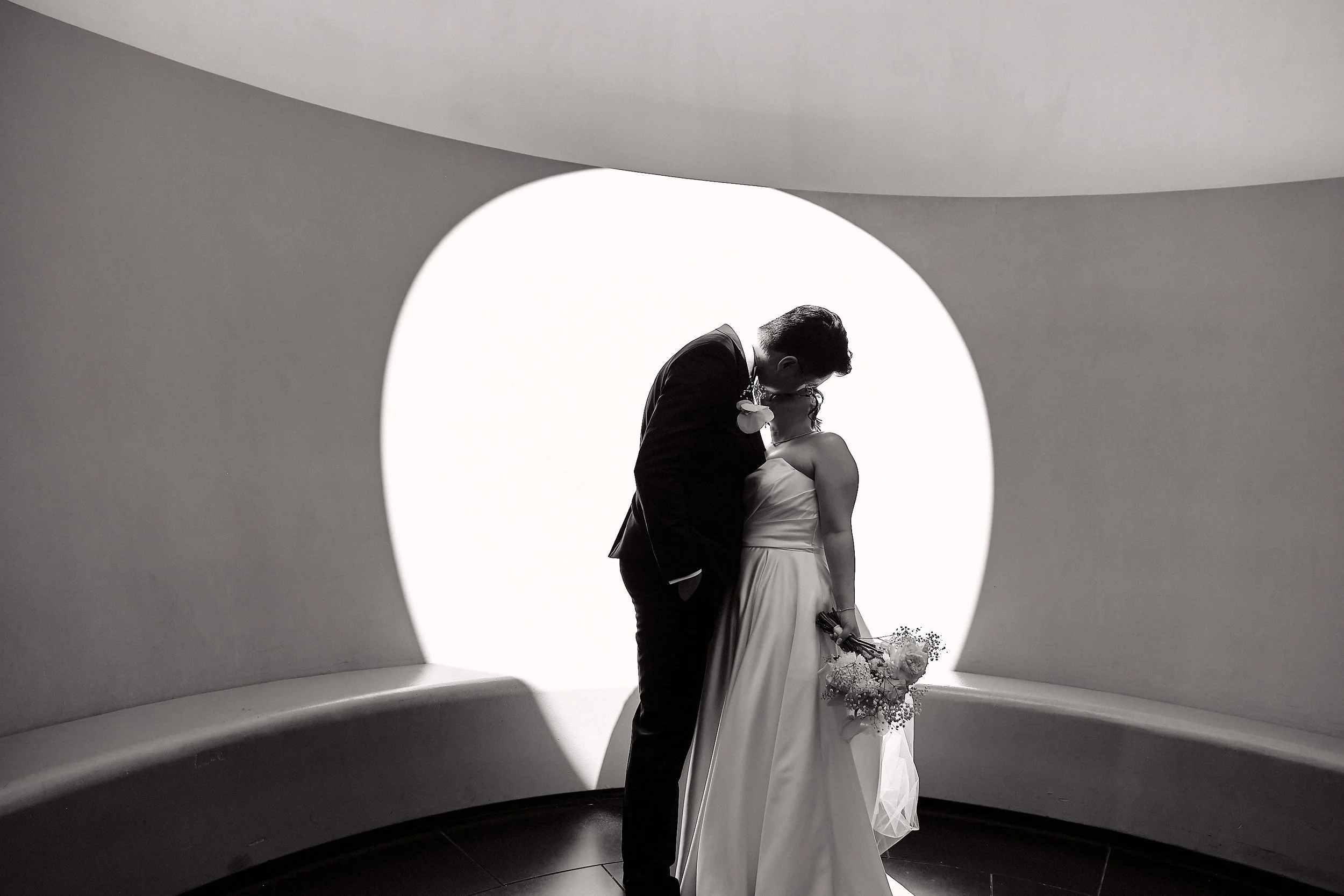 A black and white photo of a couple kissing inside a modern, curved architectural structure with a large circular opening
