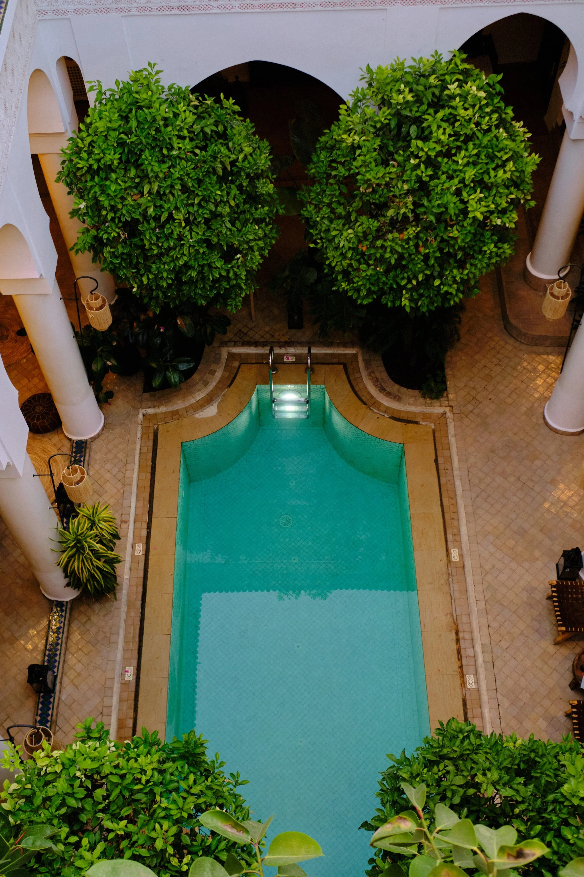 A view from above of a courtyard with a rectangular swimming pool, surrounded by lush green bushes, white columns, and tiled flooring, with decorative lamps hanging on the sides.