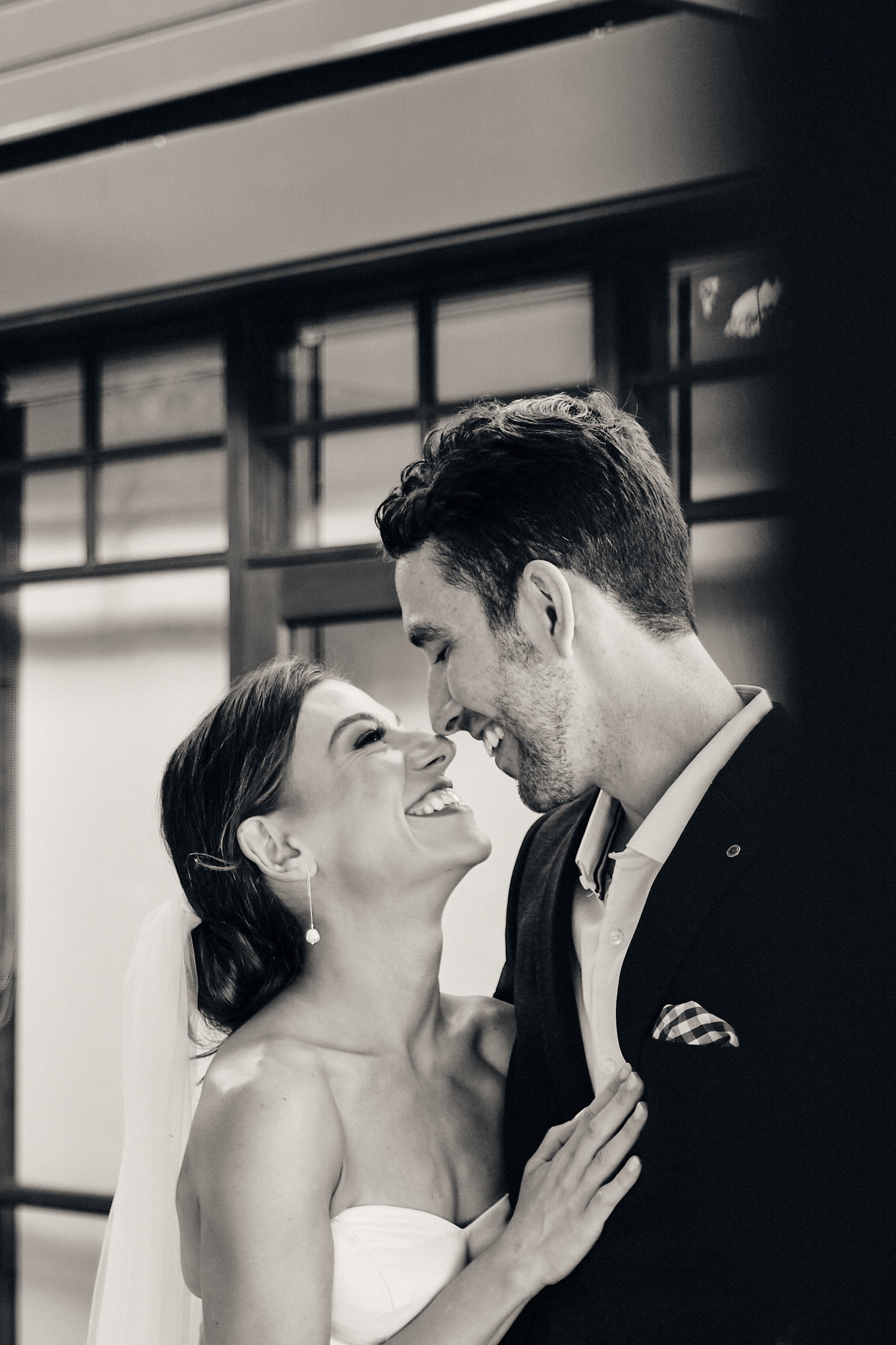 A bride and groom are close together, smiling, with their foreheads touching. The bride is in a strapless dress, wearing a veil and earrings, and has her hand on the groom's chest. The groom is in a tuxedo with a patterned pocket square, smiling back