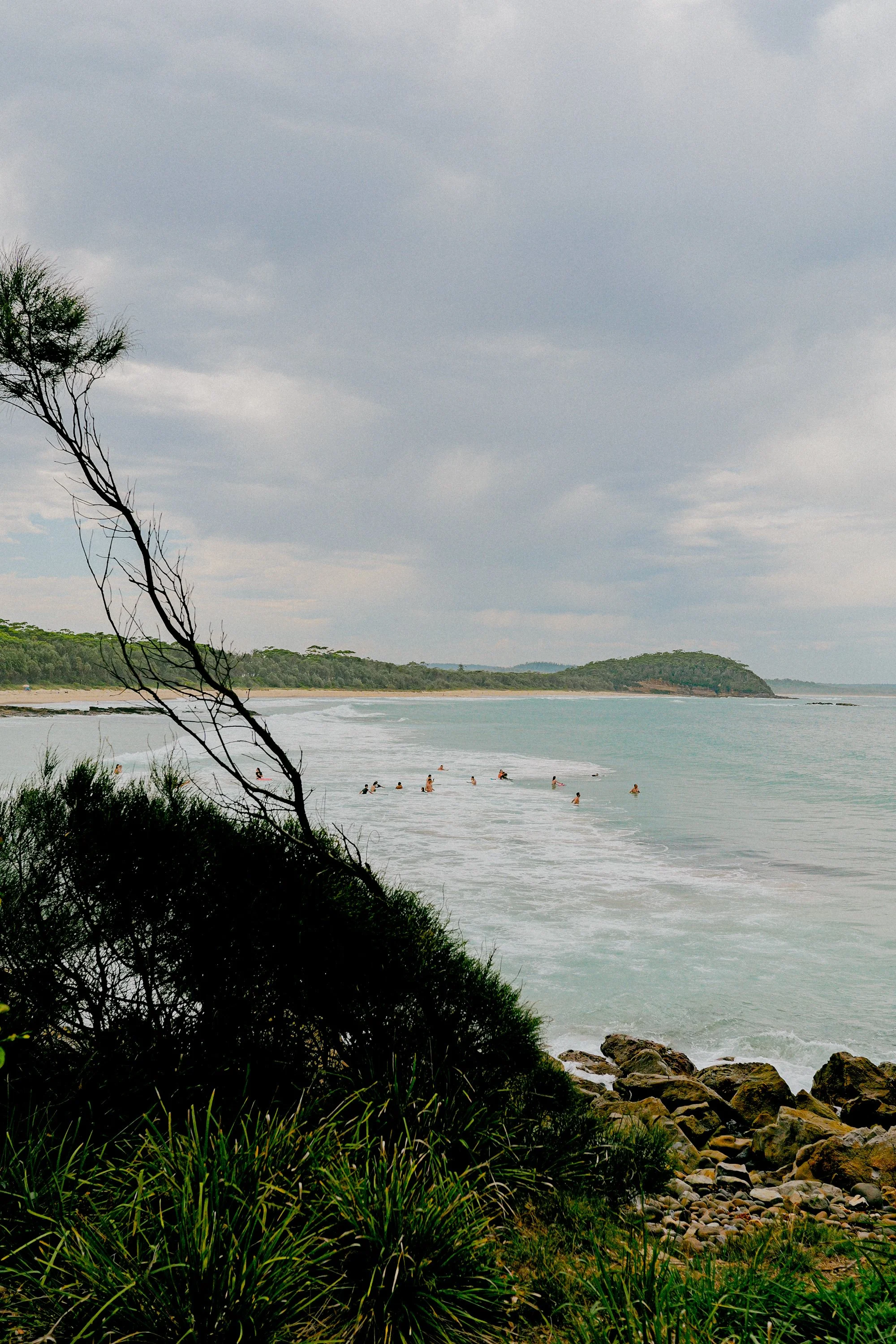 Overcast sky over a beach with a group of people swimming, surrounded by green hills, rocks, and lush vegetation in the foreground.