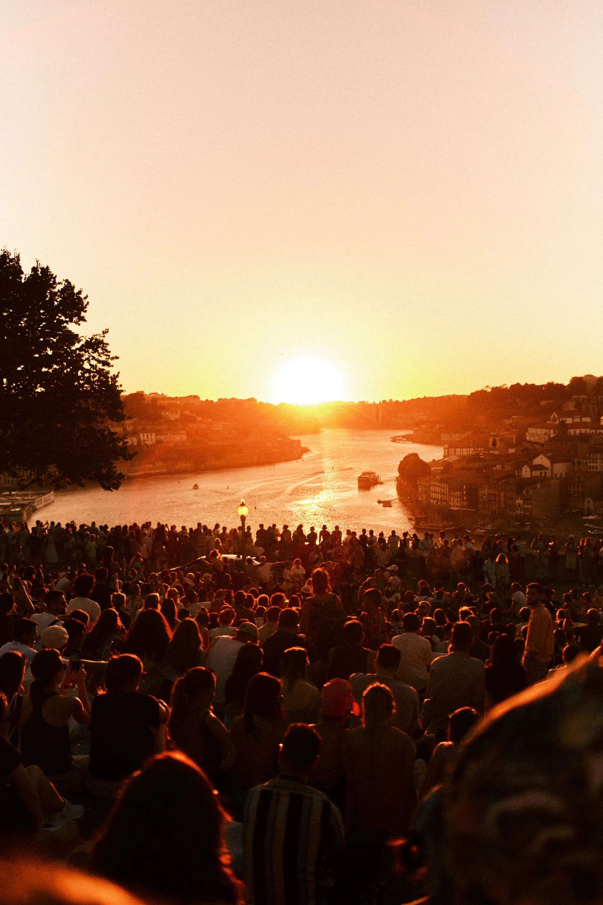 Crowd of people watching a sunset over a river with trees and buildings on the riverbanks.