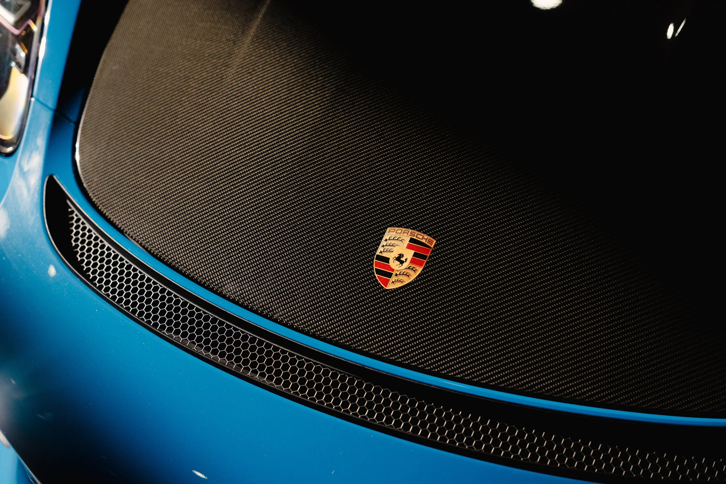 Close-up of a Porsche sports car hood, showing the Porsche emblem on a carbon fiber surface with blue exterior paint and mesh grille.