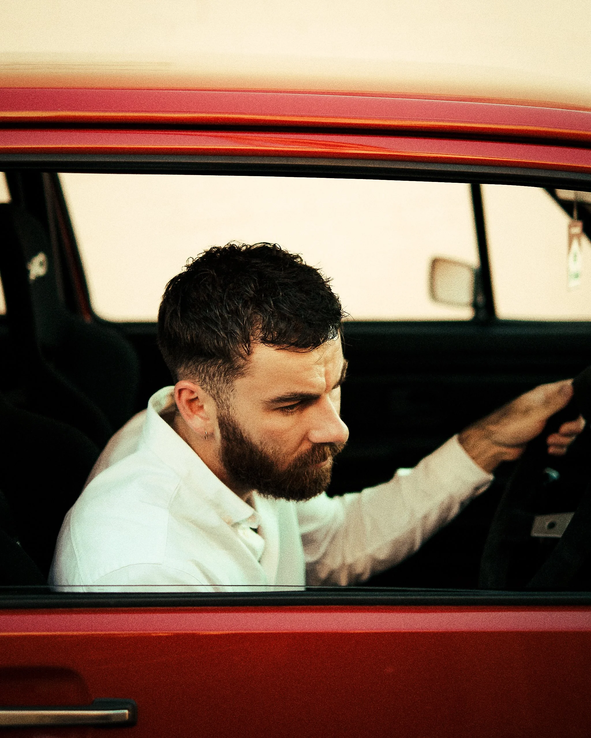 A man with dark hair, a beard, and a mustache wearing a white shirt is sitting inside a red vintage car, looking down and leaning forward.