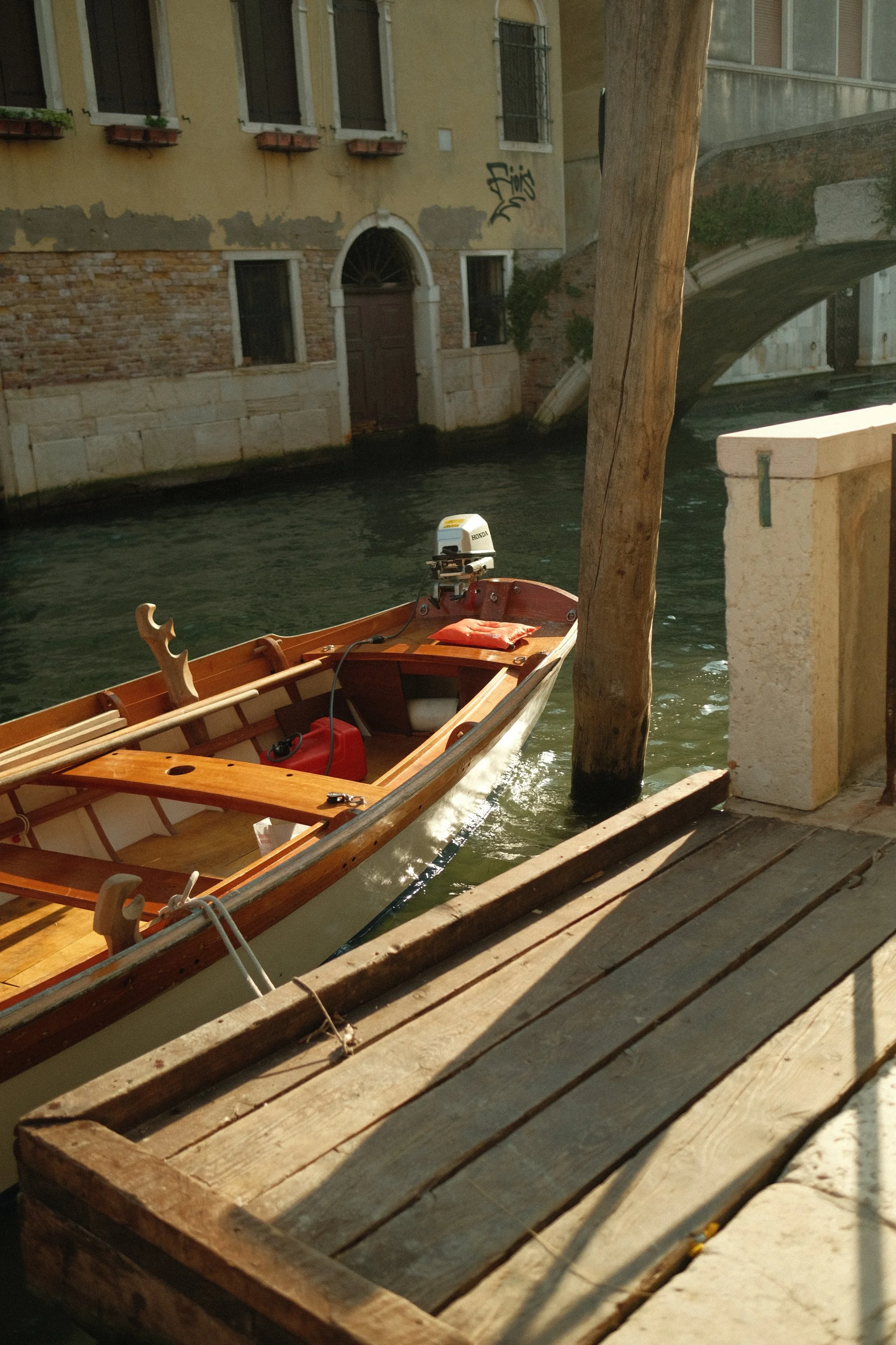 A small wooden boat with a Honda outboard motor docked near a canal, with a wooden deck and a stone wall beside it, and a building with windows and graffiti in the background.
