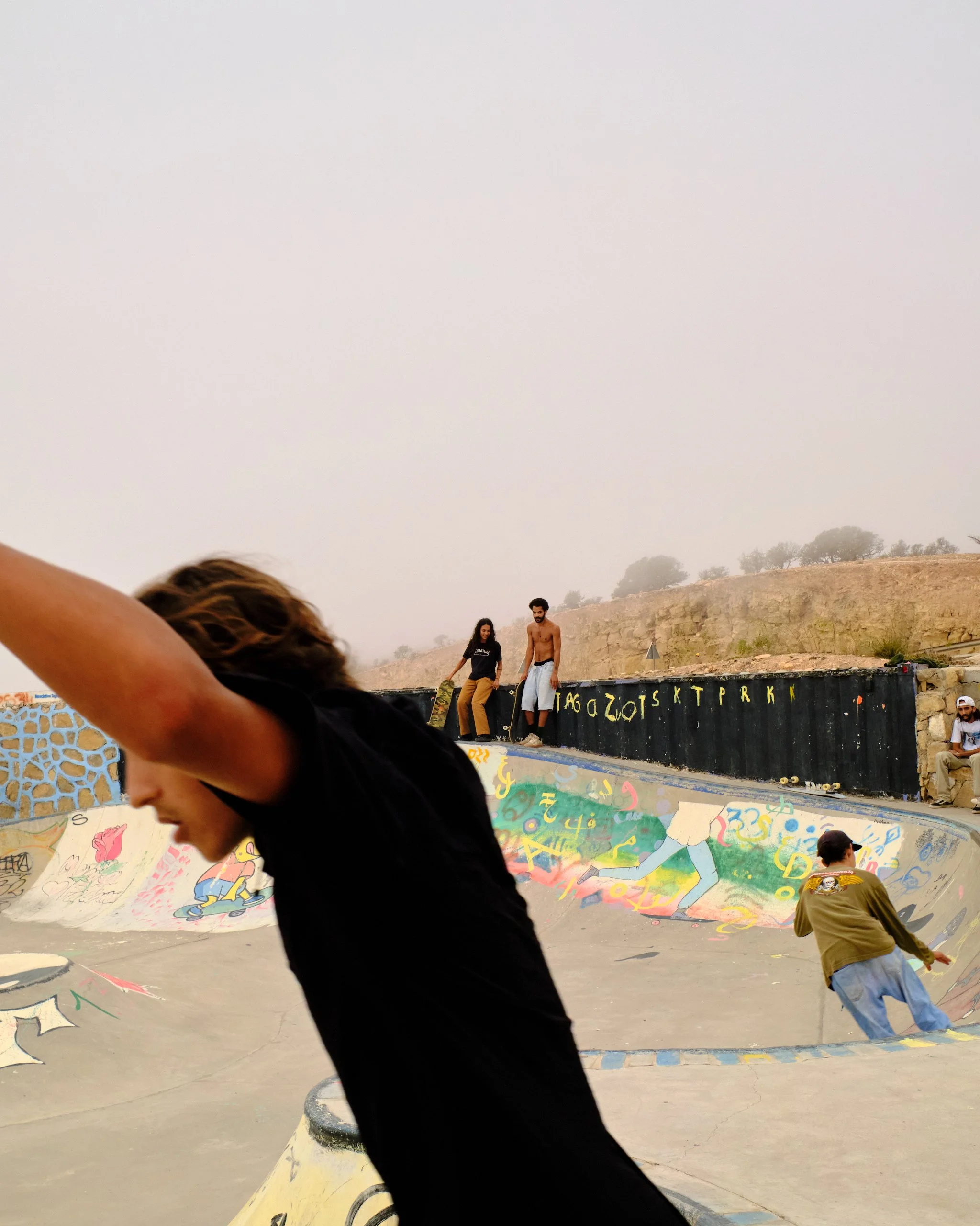 People at a skate park, with a large colorful mural on the wall, trees and hills in the background, and a cloudy sky overhead.