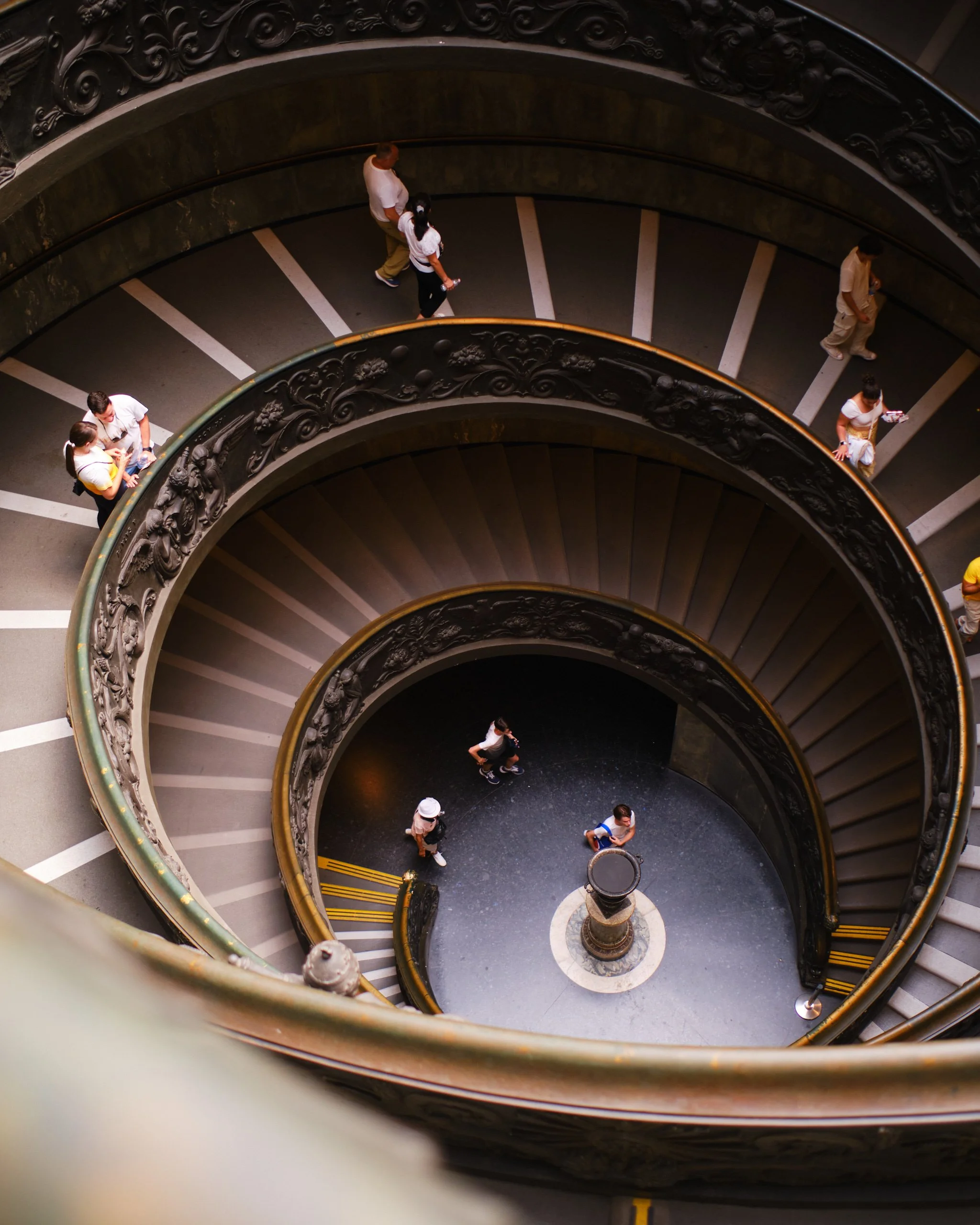A bird's-eye view of a spiral staircase with ornate black railings and a marble wall, showing several people walking on the stairs below.
