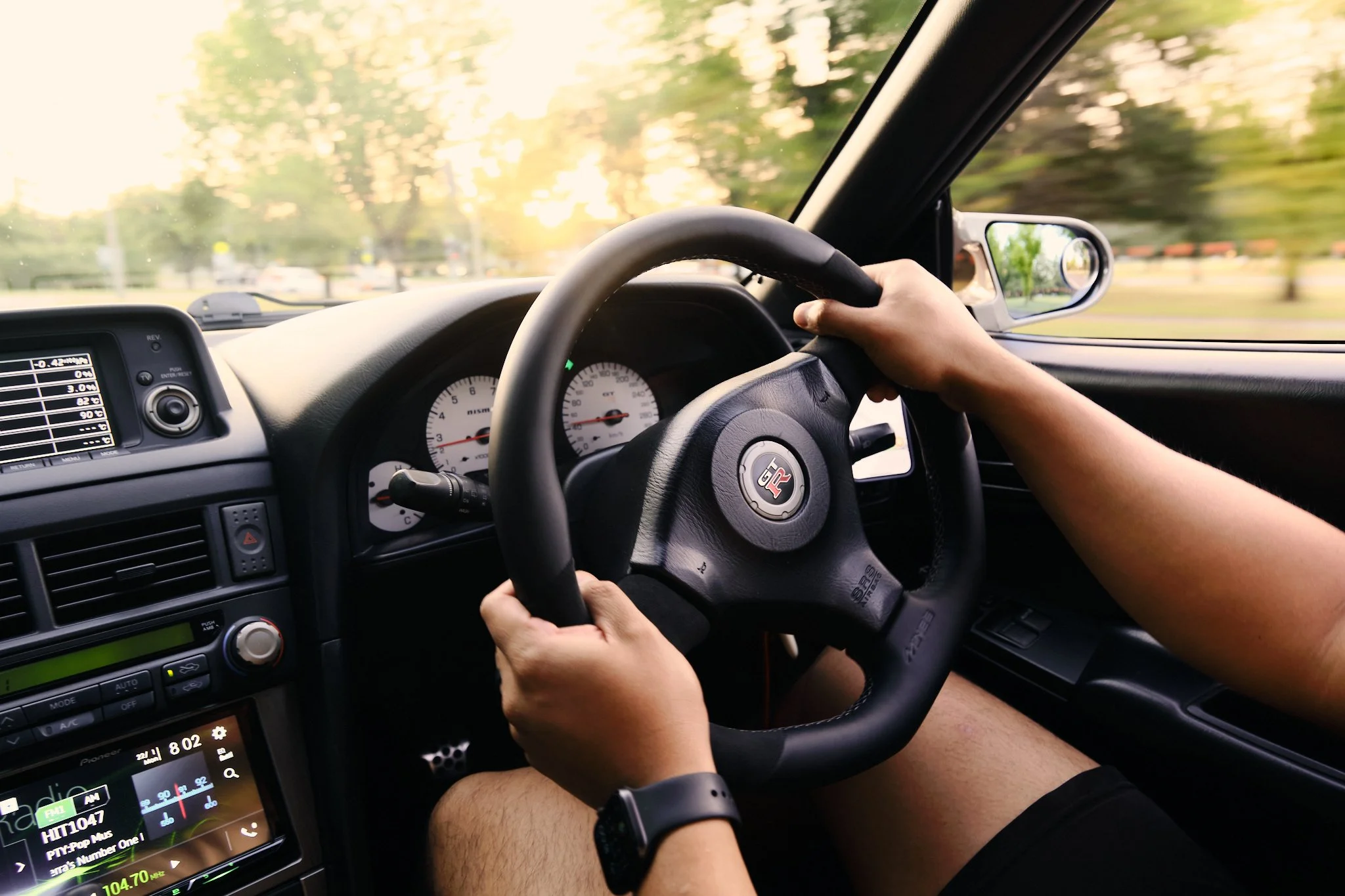 A person driving a car with a view of the dashboard, steering wheel, and side mirror, with trees and sunlight visible outside.