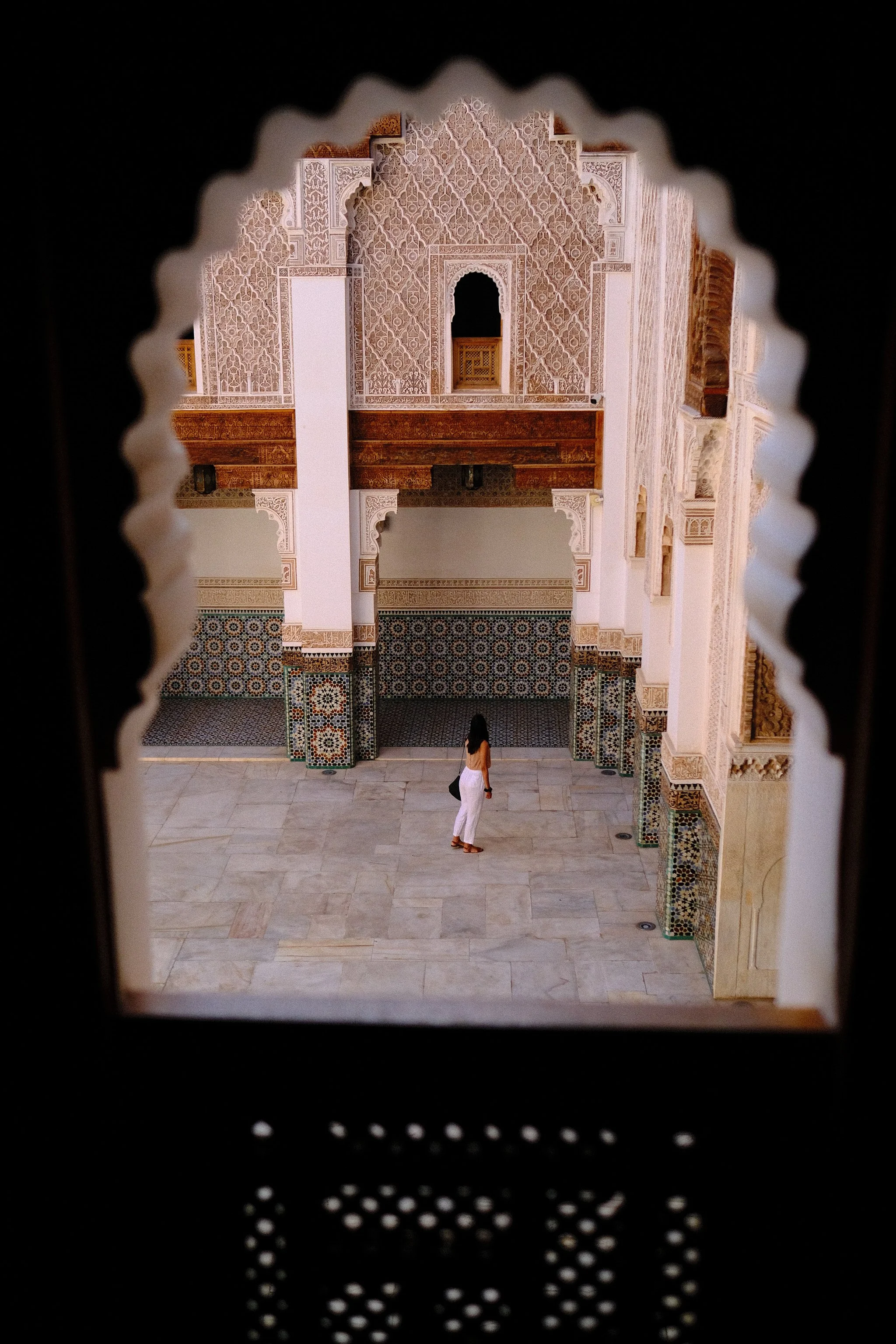 View through a decorative perforated screen showing a woman in white pants and a top walking in a traditional Moroccan courtyard with ornate tile work and intricate plasterwork.