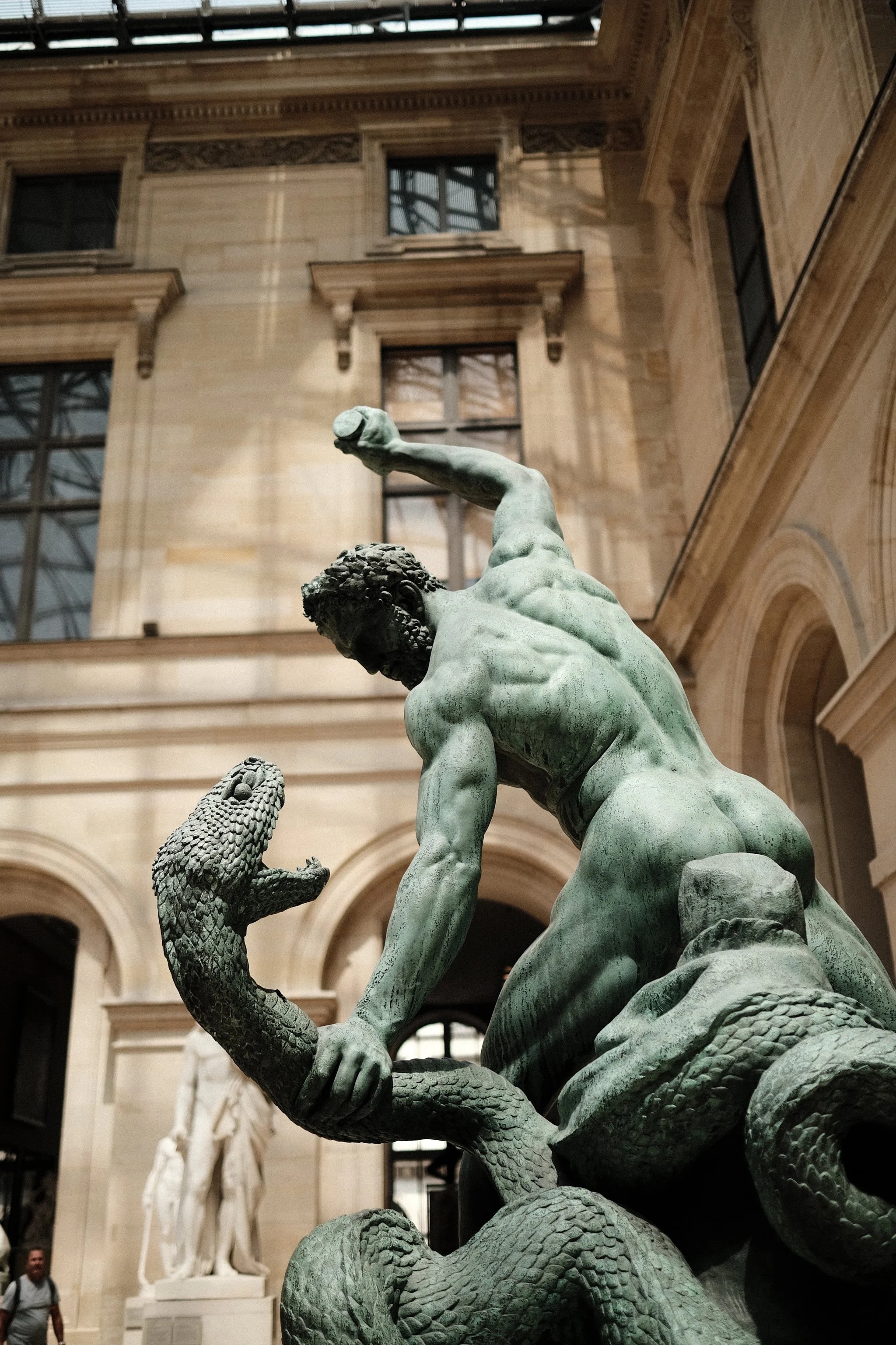 A bronze statue depicts a muscular man battling a snake, with the man's arm raised and gripping the snake's head. The scene is set in a grand hall with arched windows and detailed stone architecture.