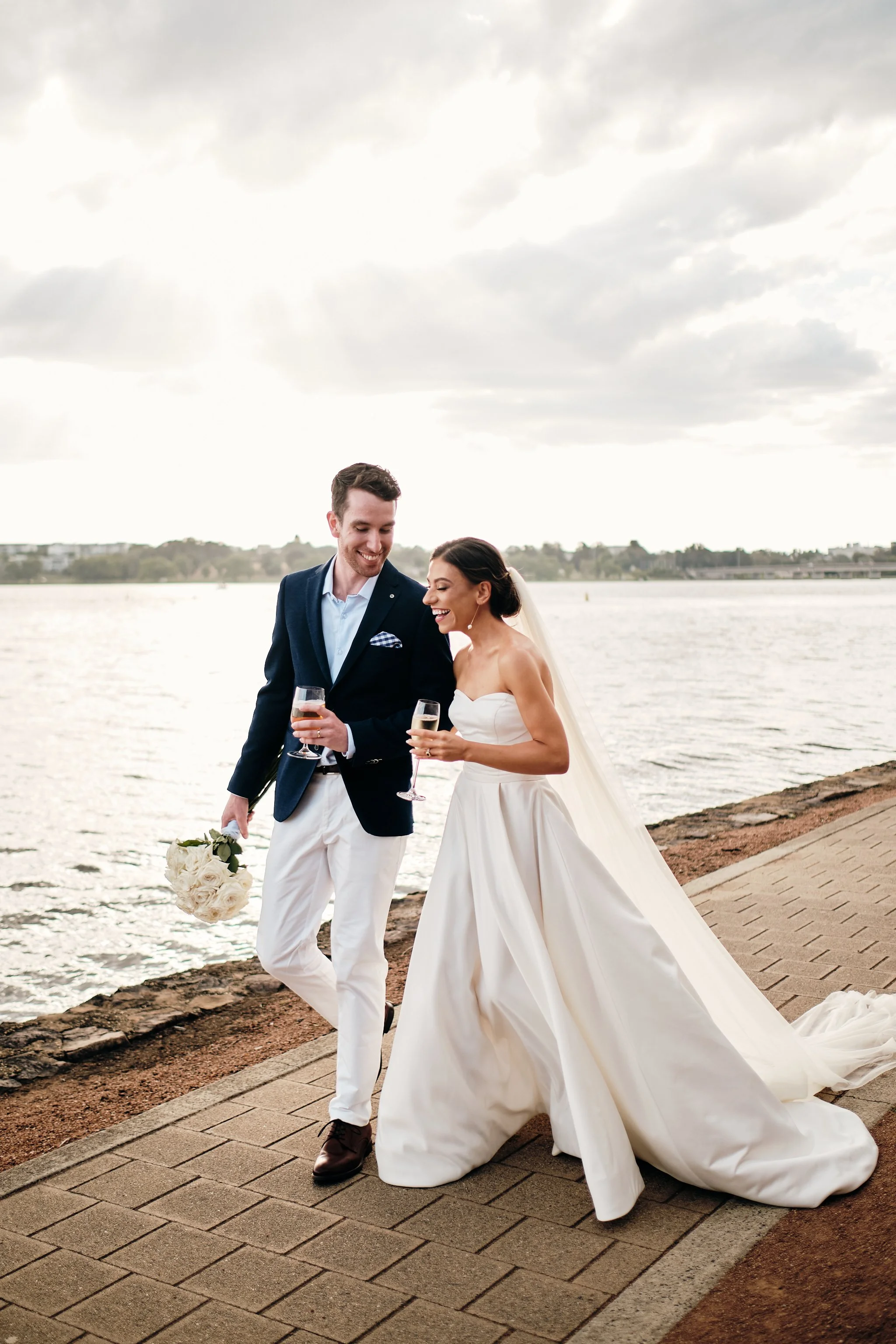 A newlywed couple walking by a body of water, smiling and holding champagne glasses, with a bride holding a bouquet of white roses, in formal wedding attire, on a paved walkway with a cloudy sky.