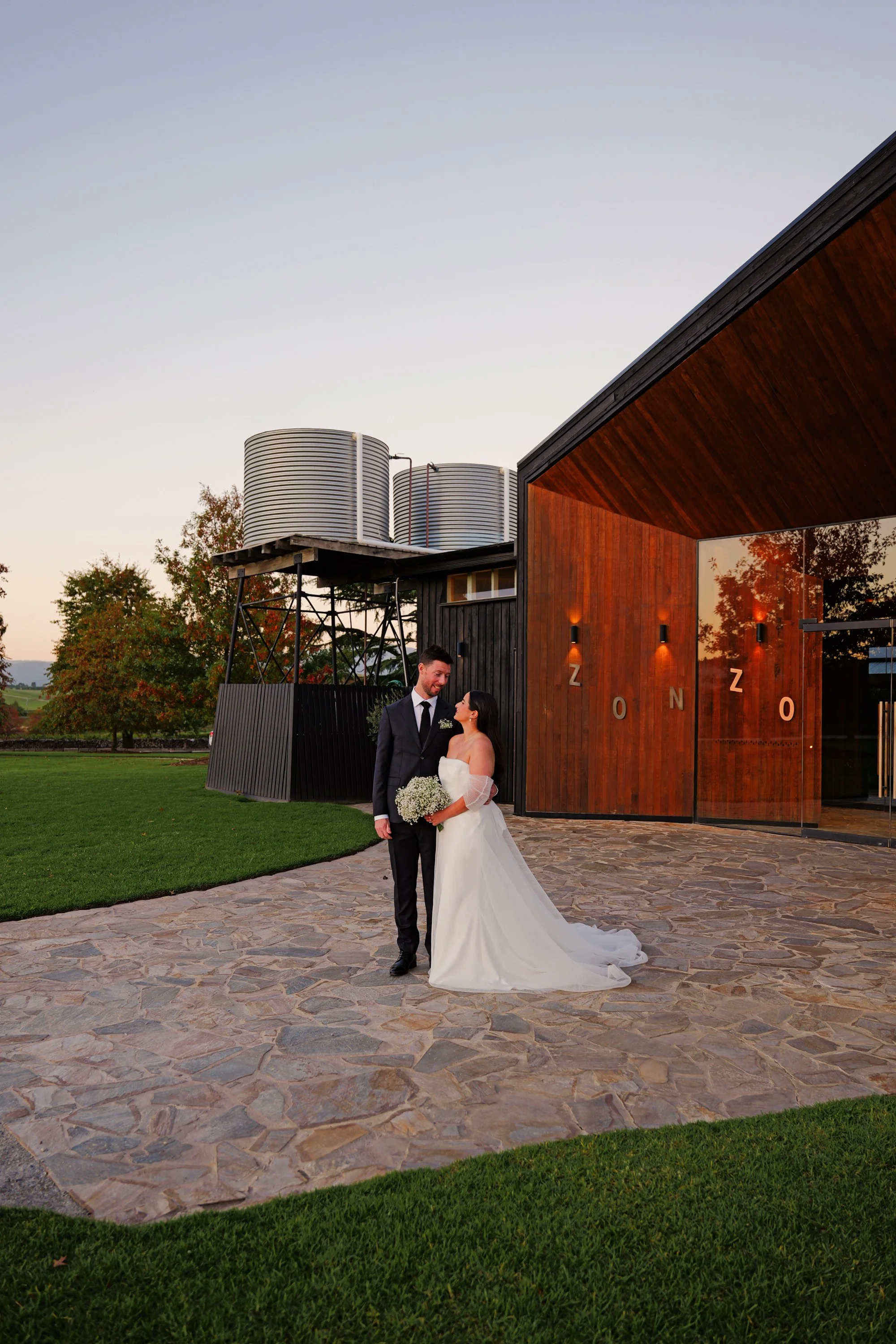 A bride and groom standing together outside a modern building with wooden walls and large glass doors, during sunset. The bride is holding a bouquet and wearing a white wedding gown, while the groom is in a dark suit.