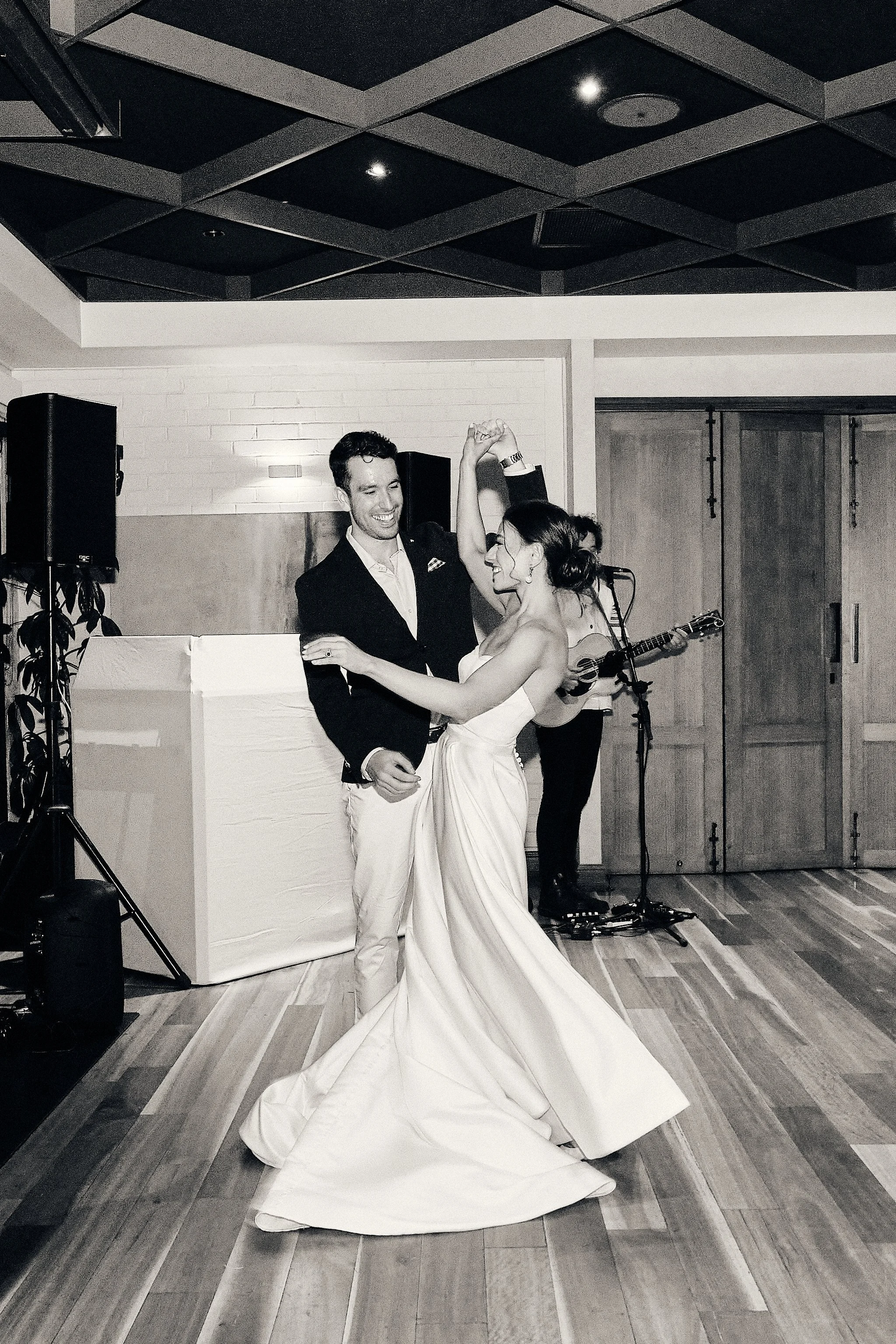 A joyful couple dancing at a wedding reception, with a musician playing guitar in the background. The bride is wearing a long, flowing gown with a train, and the groom is in a blazer and light-colored pants. The scene is in a room with a wooden floor