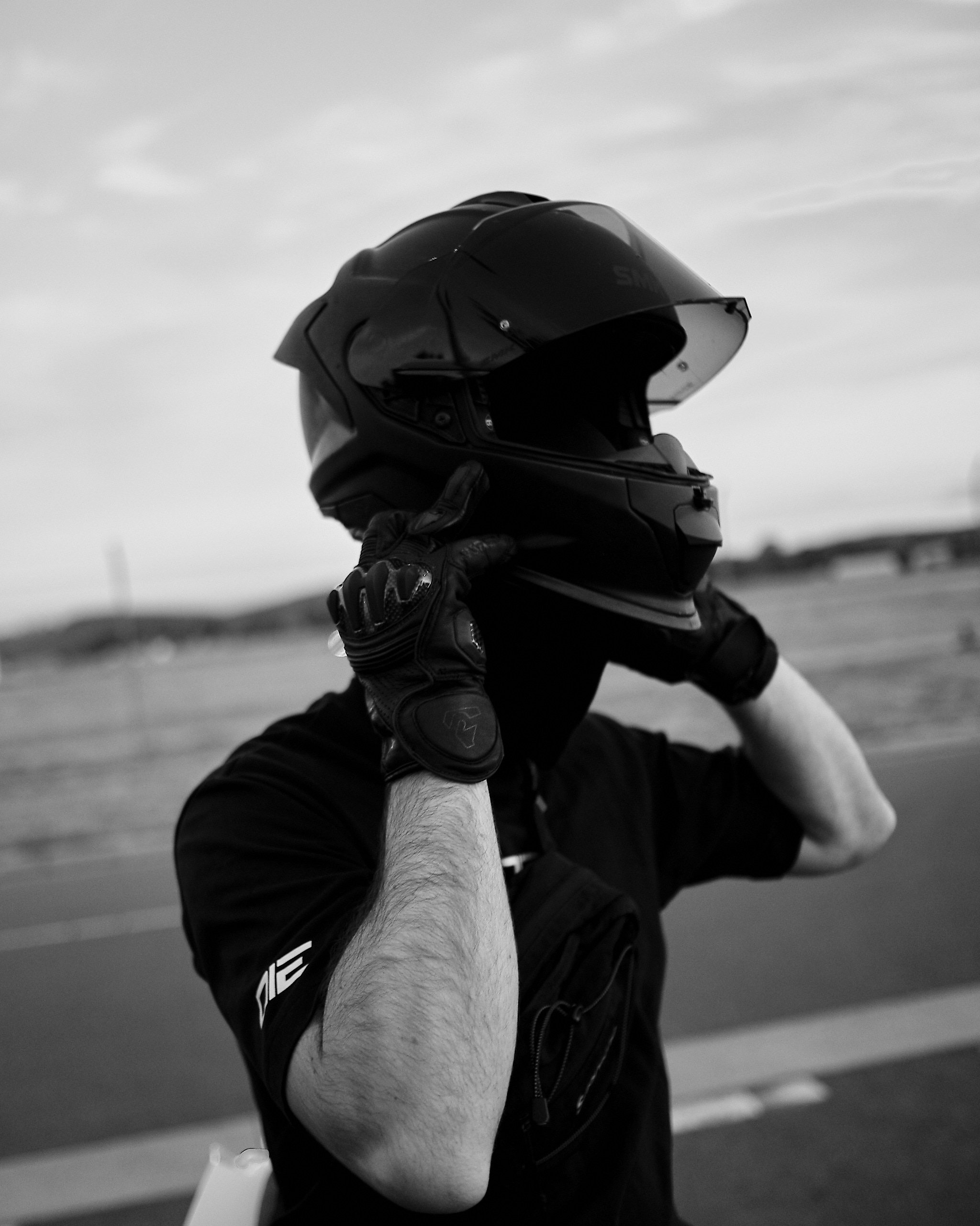 Person wearing a motorcycle helmet and gloves, holding a motorcycle helmet in front of their face, outdoors with a cloudy sky.