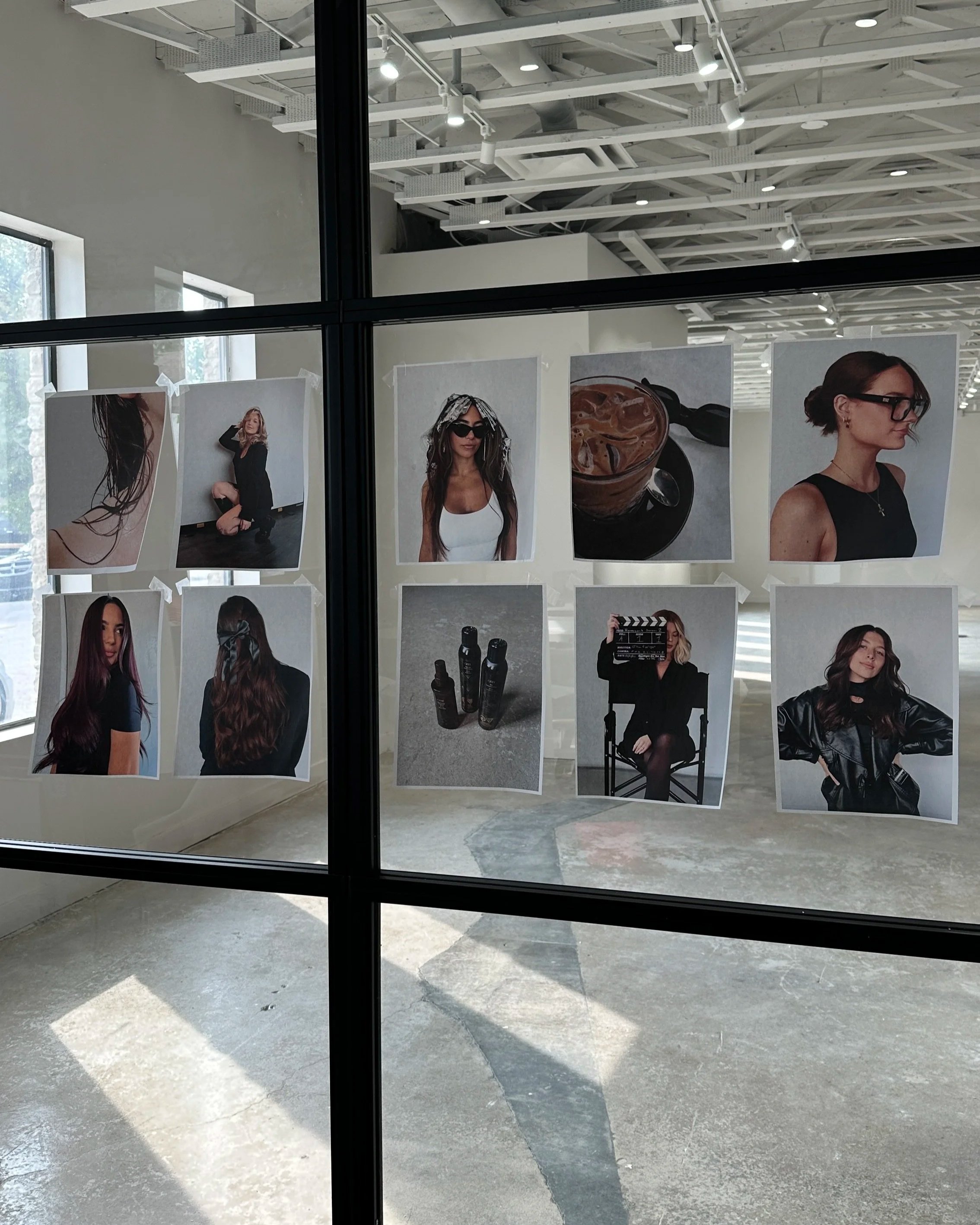 Photographs of women, hair products, and a glass of iced coffee are displayed on a glass wall in an art studio or gallery space.