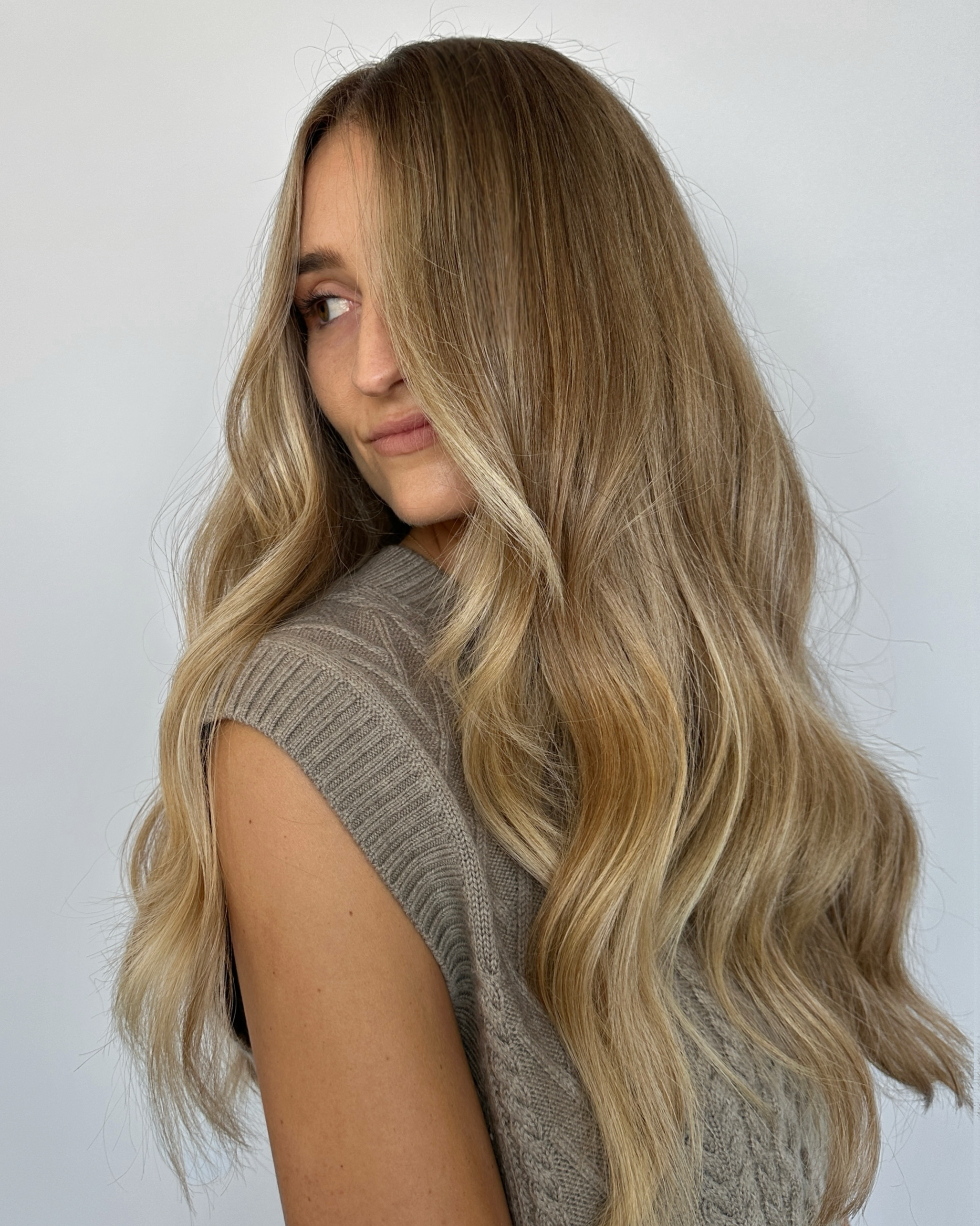 A woman with long, wavy blonde hair looking to her left, wearing a sleeveless beige sweater against a plain white background.