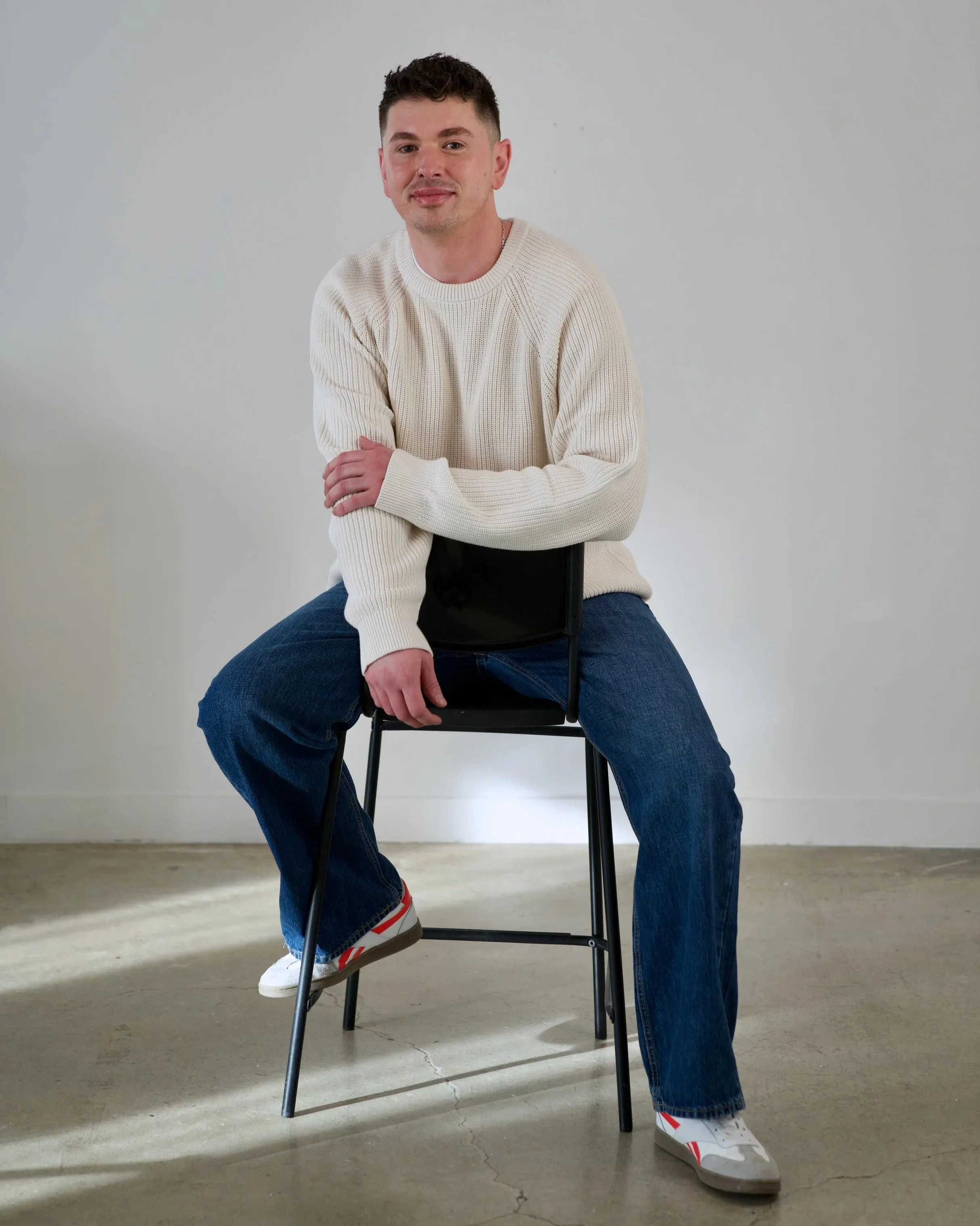 Young man with dark, curly hair and light skin sitting on a black chair, wearing a cream-colored sweater and blue jeans, with one arm resting on the seat back, posed against a plain light gray background.