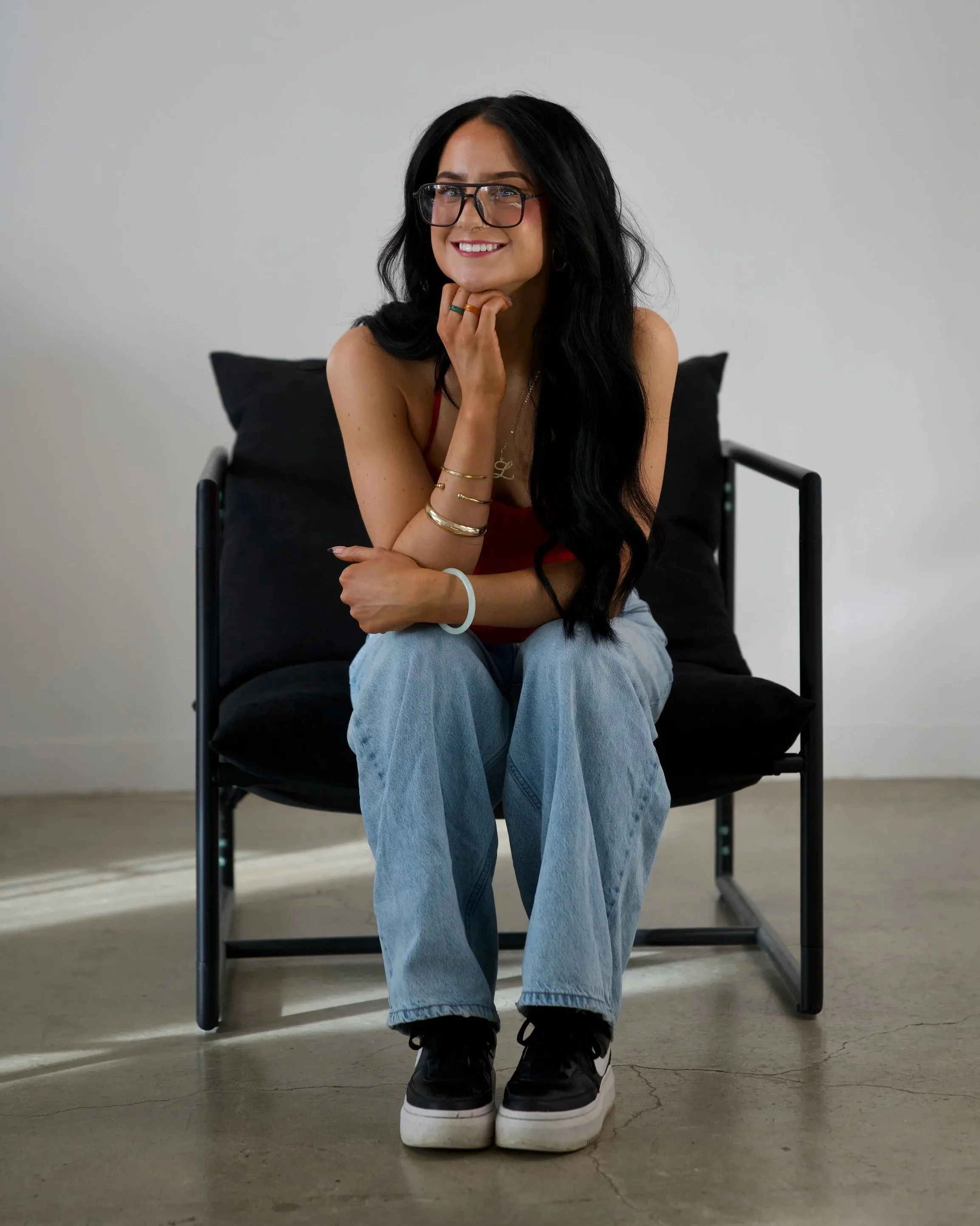 A young woman with black hair wearing a strapless black top, gold jewelry, and pink blush, touching her ear, looking to the side against a plain gray background.