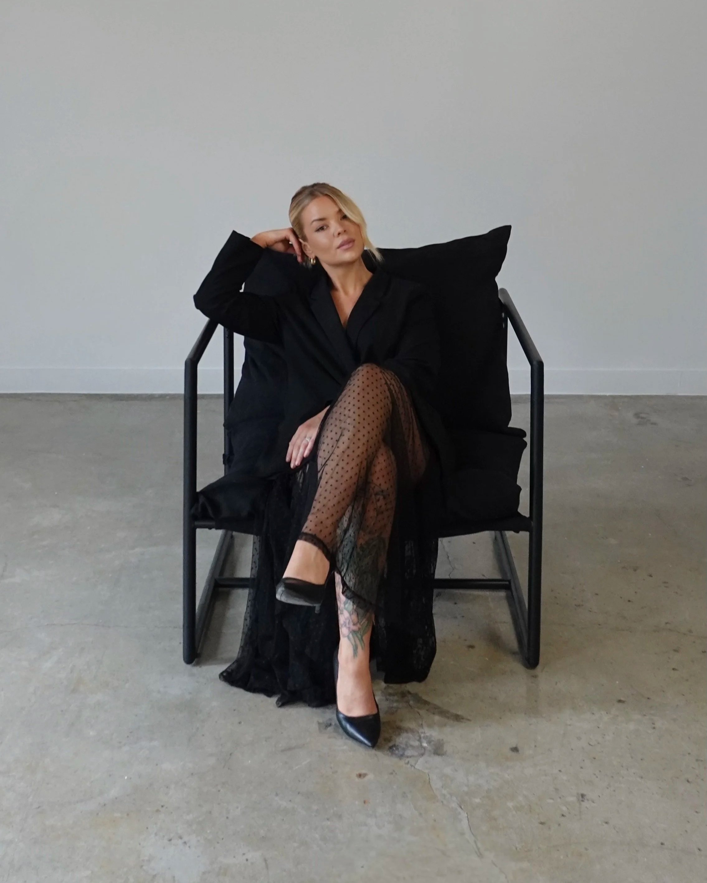 Woman sitting on a modern black chair against a plain light-colored wall, wearing a black dress and patterned fishnet stockings, with her hand resting behind her head and looking at the camera.