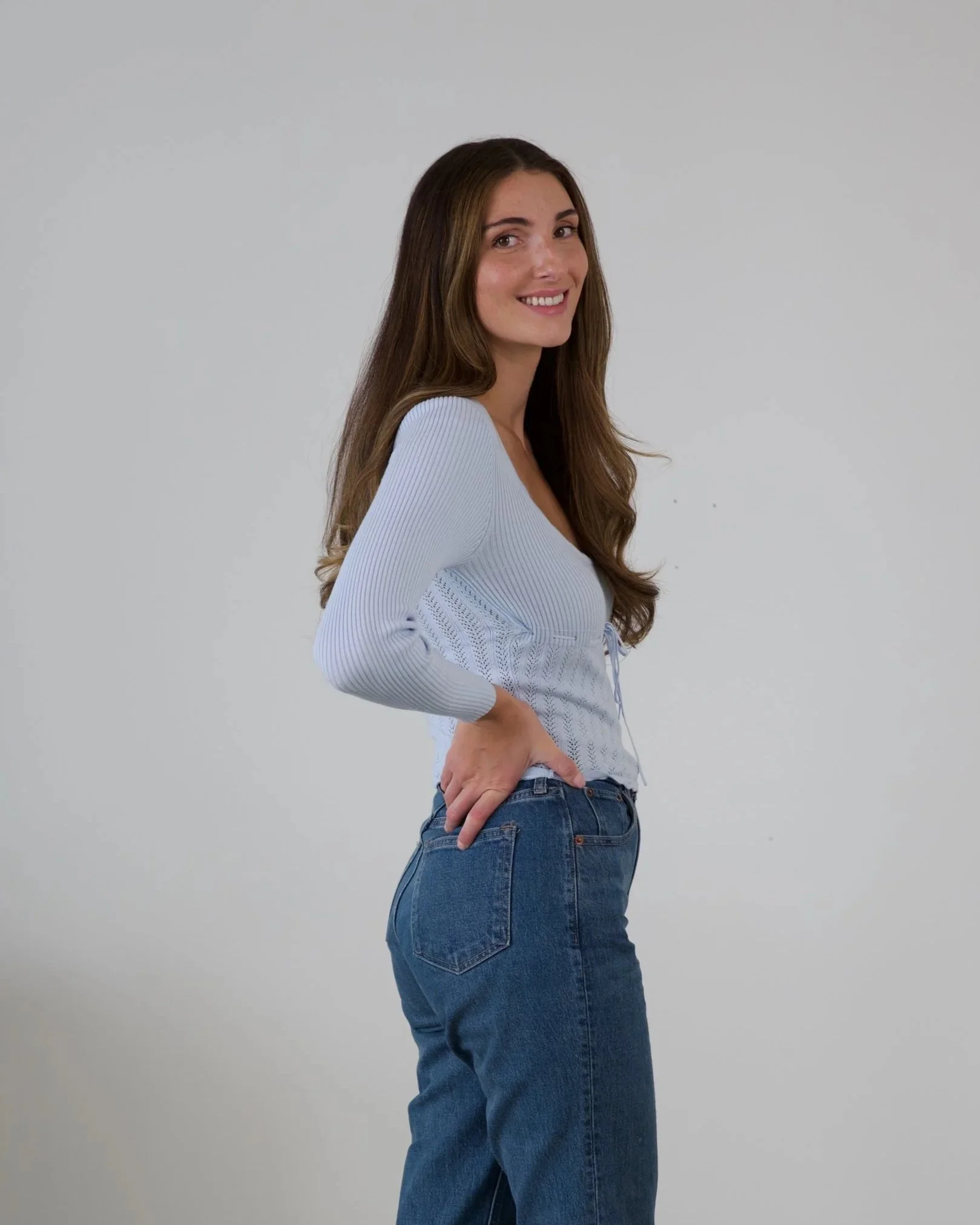 Woman sitting on a black chair with black pillow, wearing a white blouse, light blue jeans, and black high heels, in a minimalistic room.