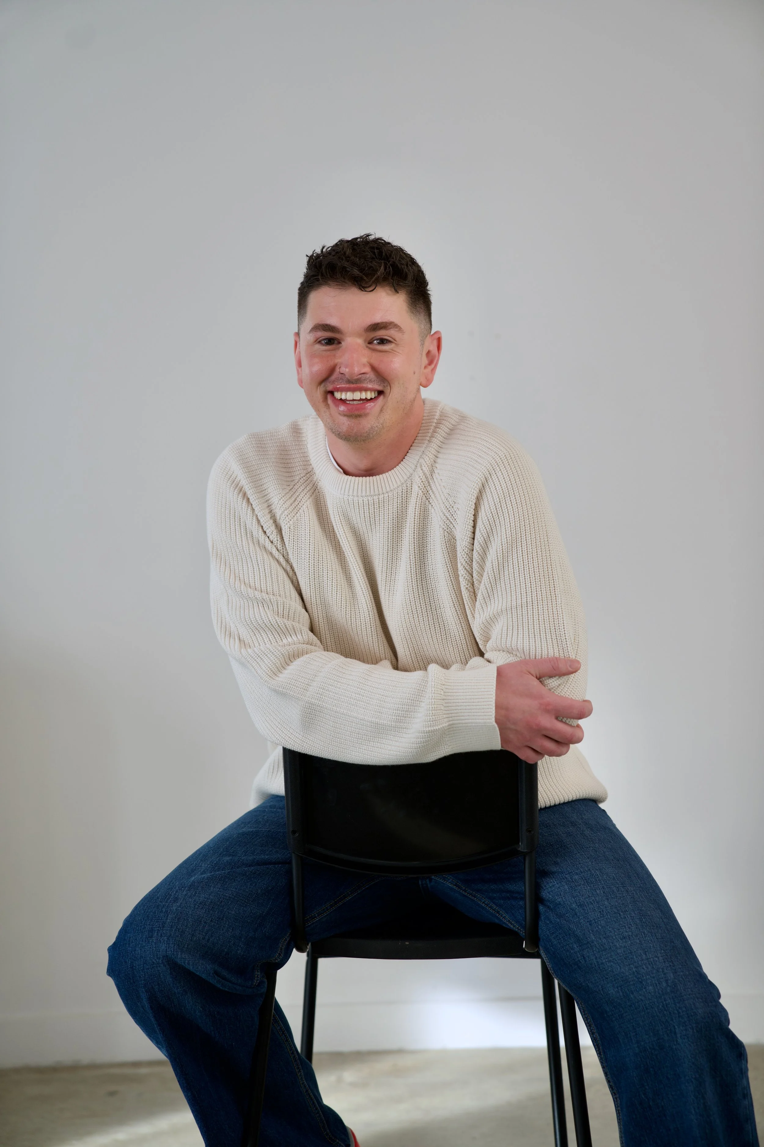 A man with dark hair and tattoos, dressed in black, sitting on a black chair with a black pillow, in a room with a plain white wall and concrete floor.