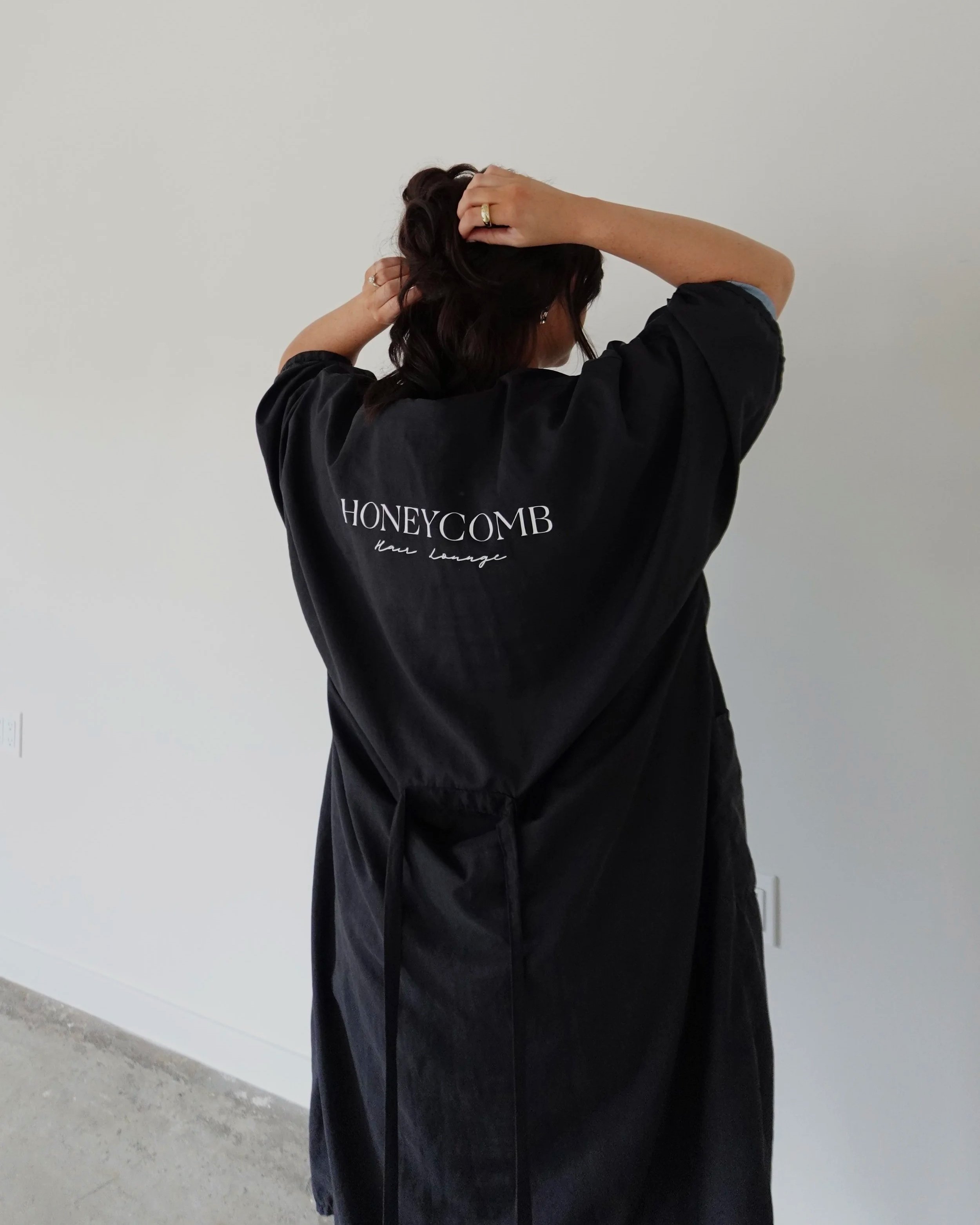 A woman with dark hair is standing against a plain white wall, adjusting her hair with both hands. She is wearing a black shirt with the words 'HONEYCOMB Hair Lounge' printed on the back.