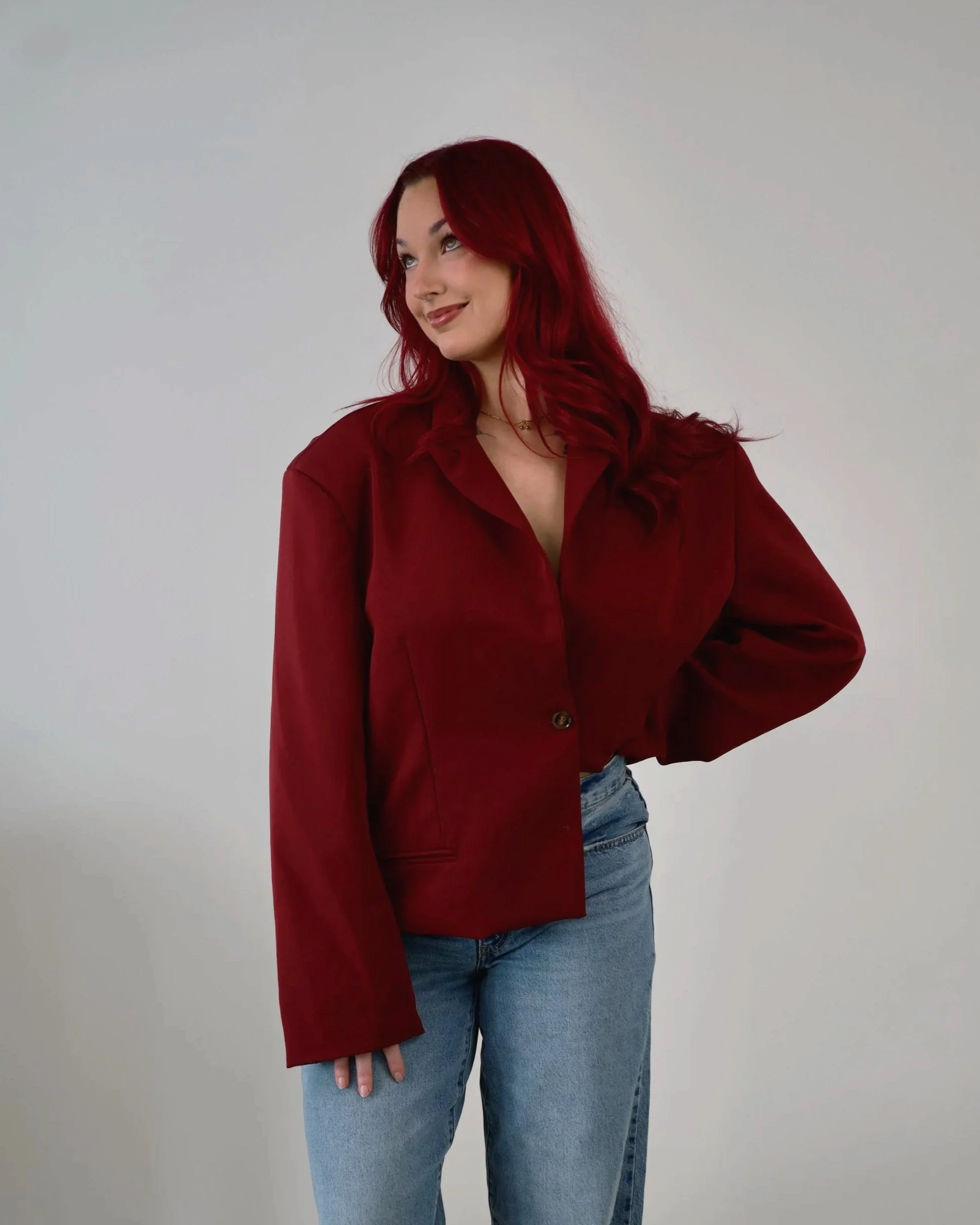 A woman with long red hair wearing a red blazer and light blue jeans, standing against a plain white background.