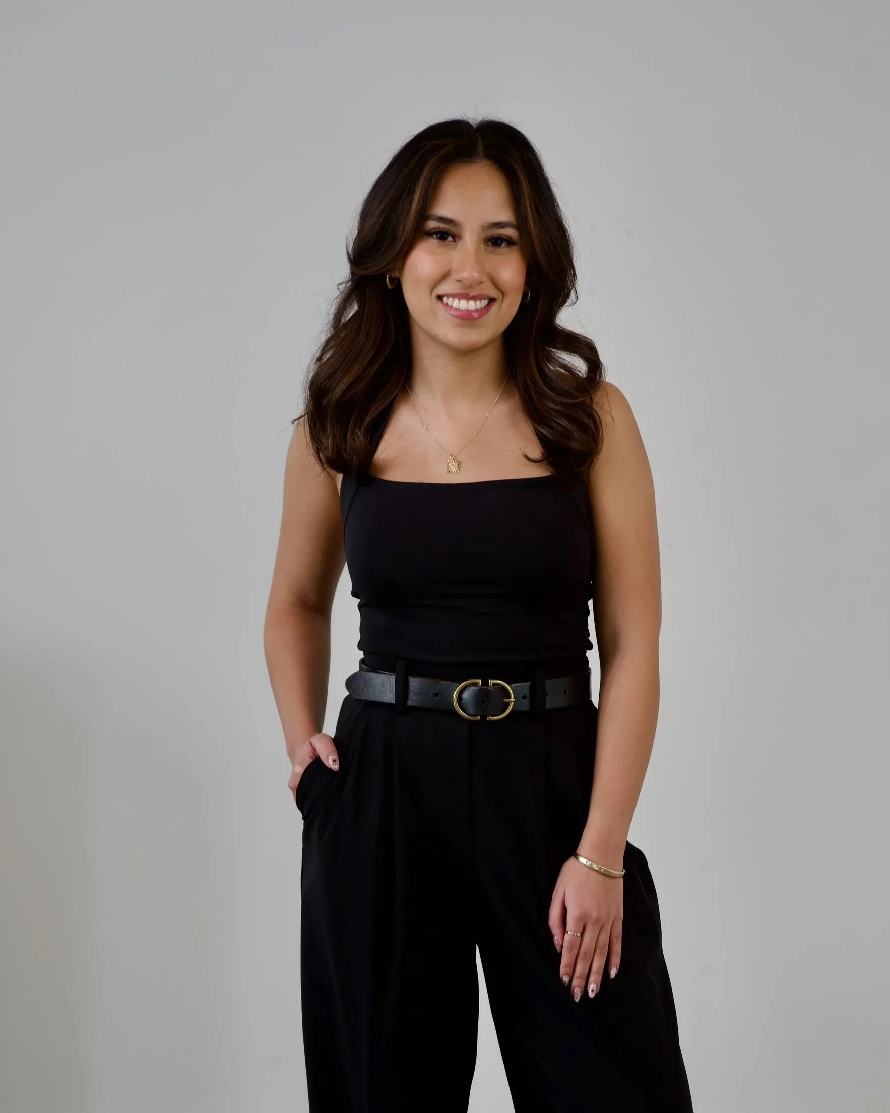 A woman with brown hair wearing a black sleeveless top and blue jeans, sitting on a black chair, smiling and touching her hair, against a plain light-colored background.