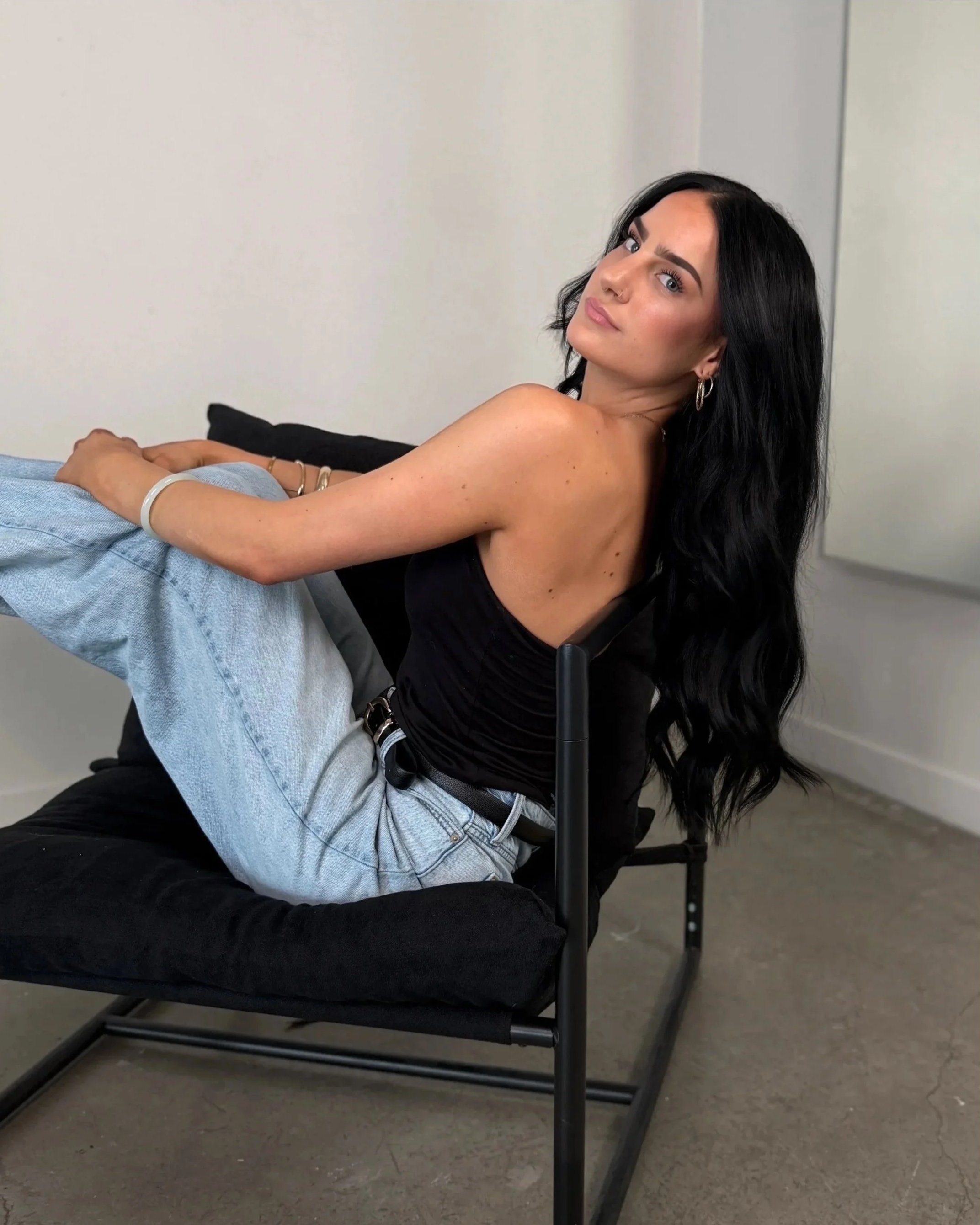 A woman with long black hair, wearing hoop earrings, resting her head on a chair, looking at the camera with a serious expression. She is wearing a black top, and there is a person next to her wearing a shirt and jeans, with their arm resting on the chair. The background shows a plain wall and the floor is concrete.