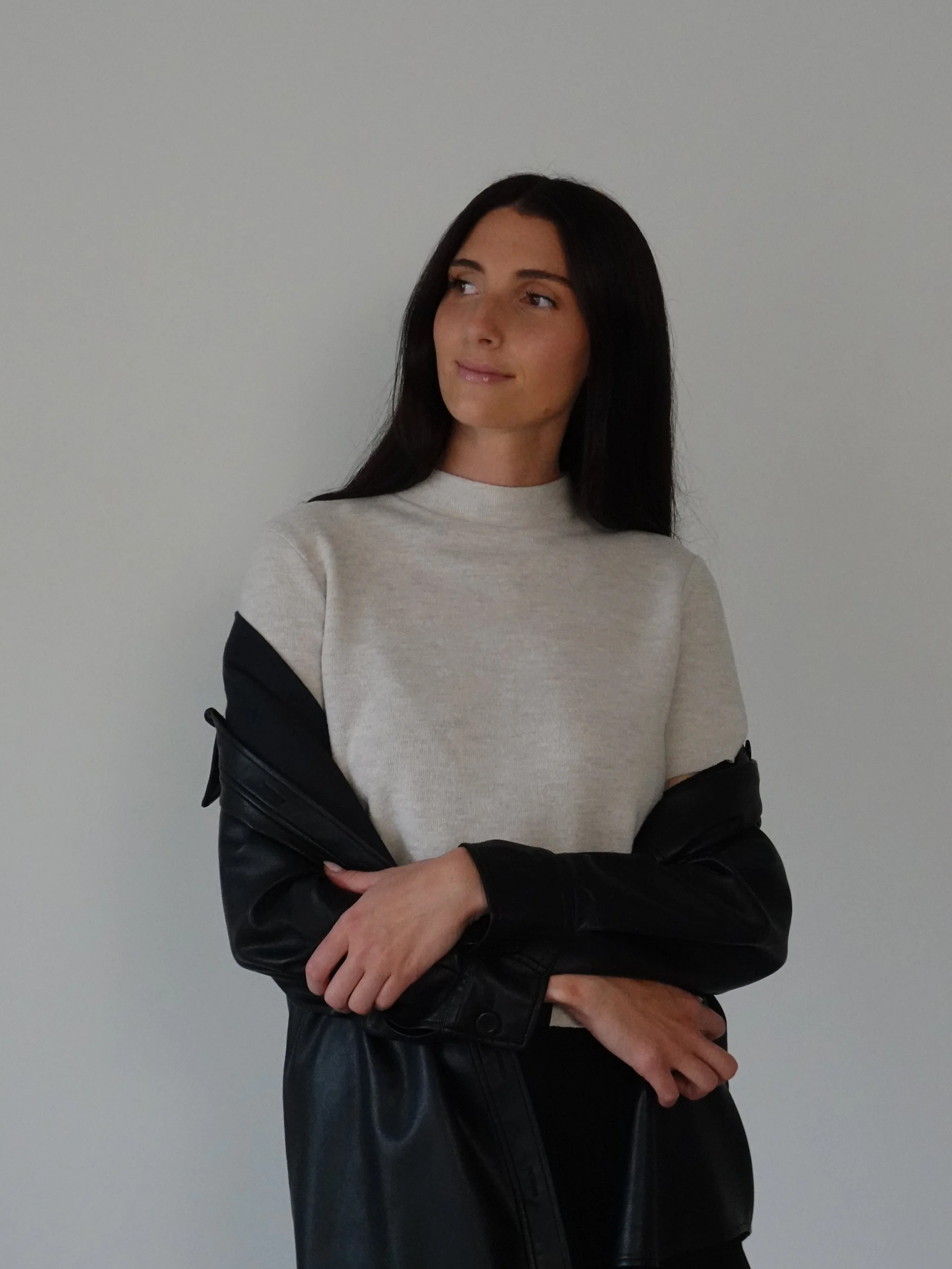 Woman sitting on a black chair with black pillow, wearing a white blouse, light blue jeans, and black high heels, in a minimalistic room.