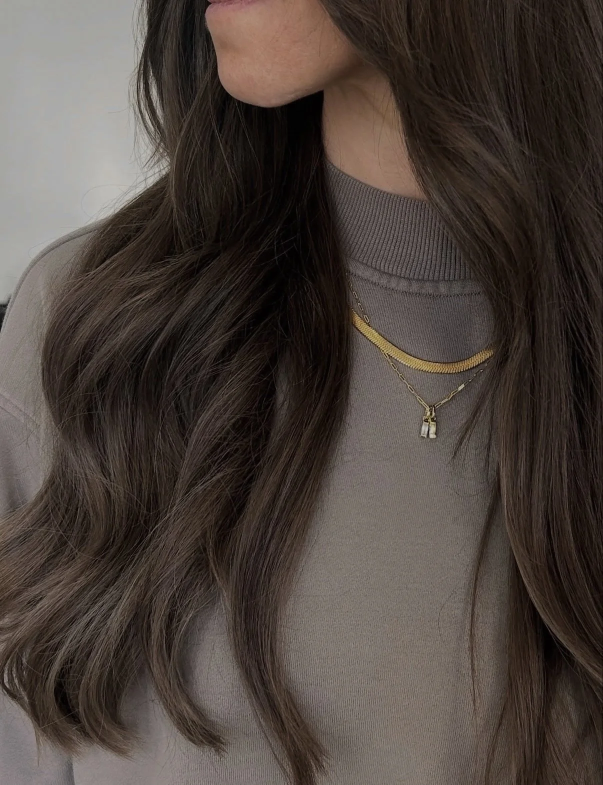A woman with long, wavy brown hair wearing layered gold necklaces and a beige top.