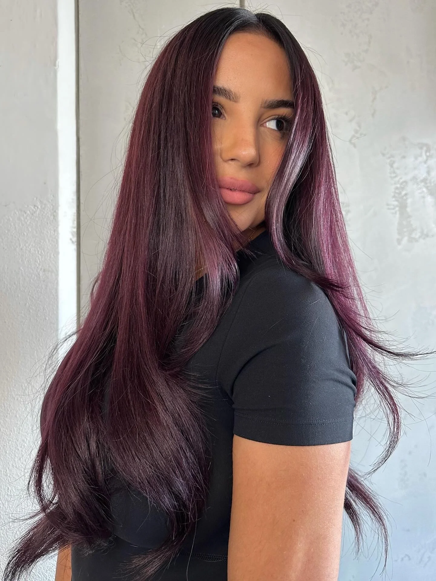 A woman with long, wavy purple hair, wearing a black shirt, looking over her shoulder.