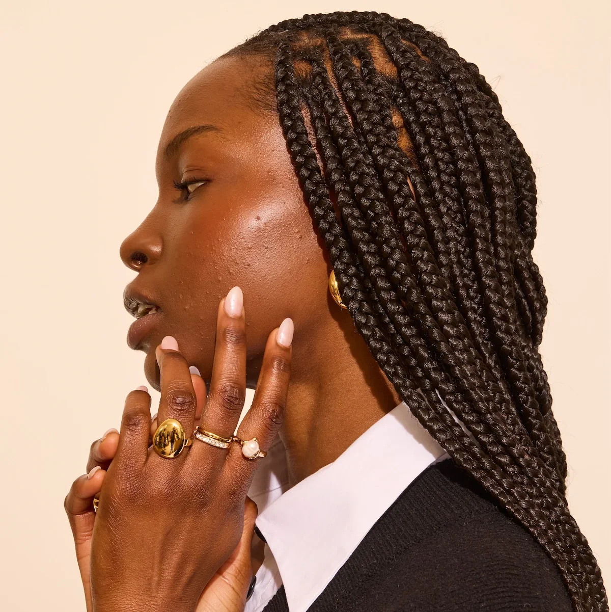 Close-up of a woman with braided hair and gold jewelry, touching her face thoughtfully, against a neutral background.