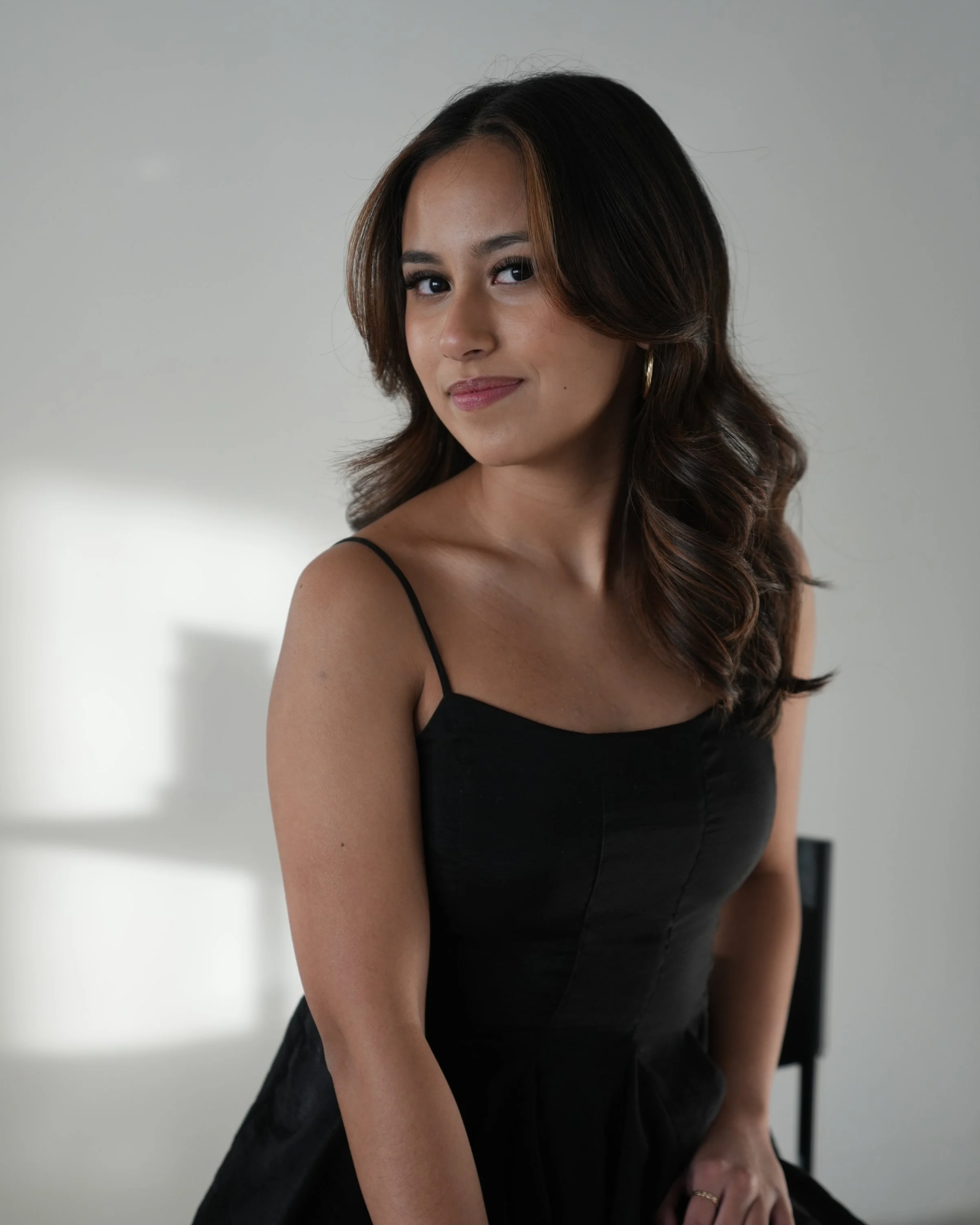 A woman with long, wavy brown hair and hoop earrings wearing a black spaghetti strap top, sitting on a chair with a neutral expression against a plain light gray background.