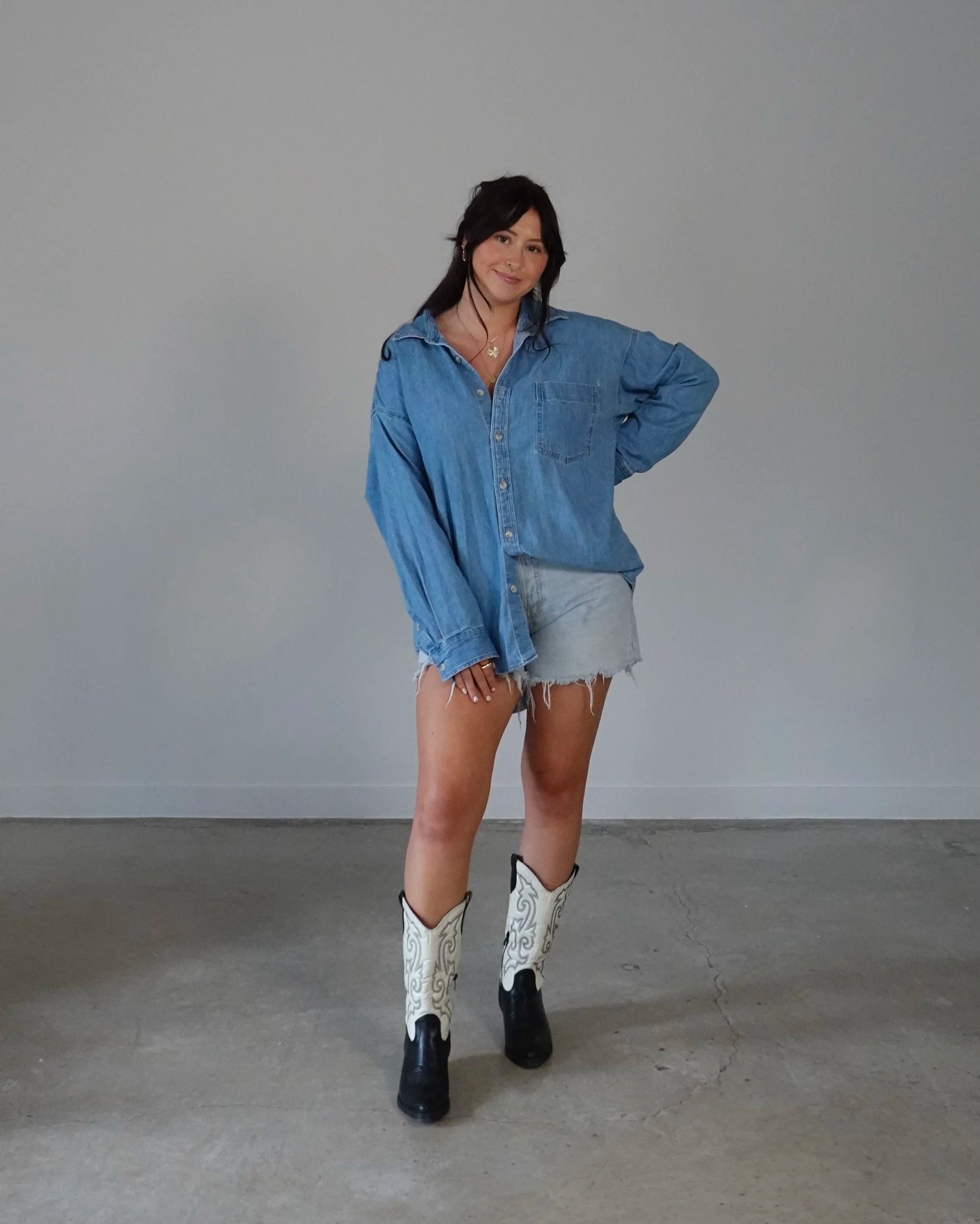 A woman with dark hair, wearing a denim shirt, gray shorts, and cowboy boots, stands against a plain gray wall with one hand on her hip and smiling.