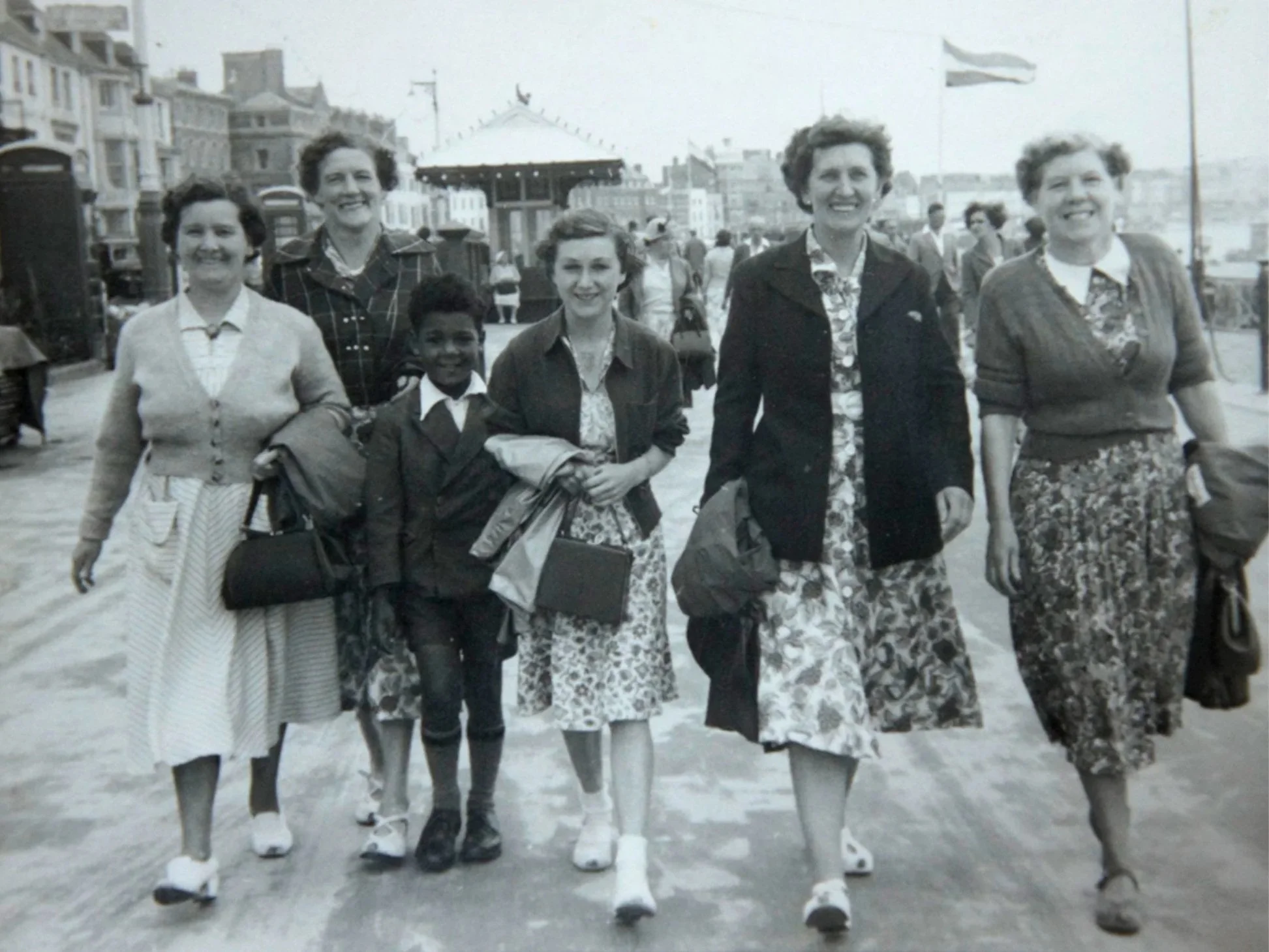 Seven women and one young girl walking along a boardwalk near the beach, some carrying bags and jackets, with a carousel, buildings, and a flag in the background.