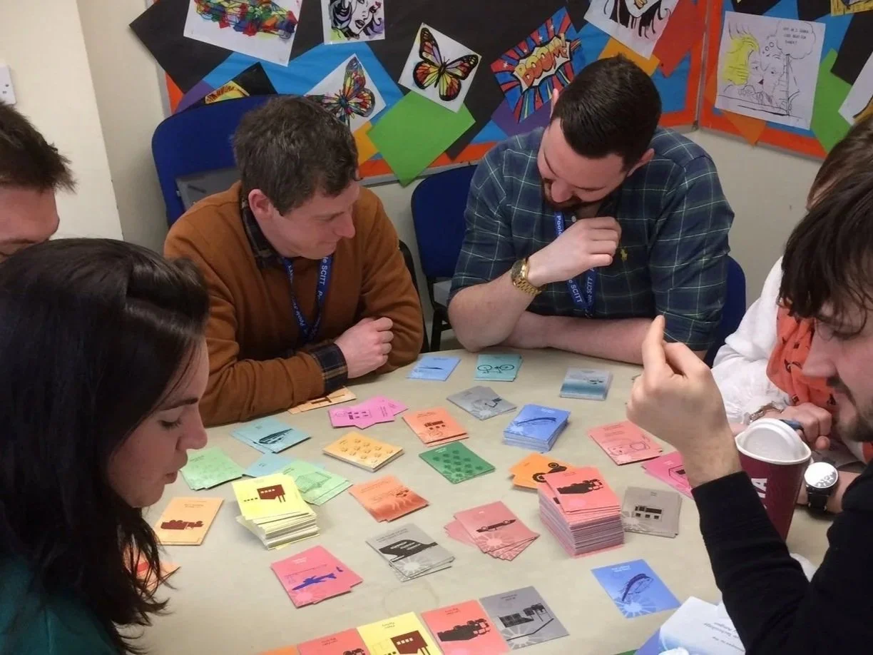 Group of people playing a card game around a table. Colorful cards with different pictures and multiple stacks of cards are on the table. The background has a bulletin board with butterfly and comic-style artwork.
