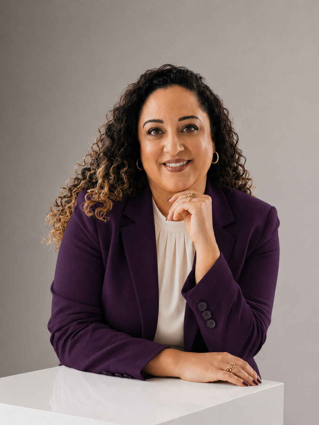 A woman with curly hair wearing a purple blazer and white blouse, sitting at a white table with her hand resting on her chin, smiling, with a neutral gray background.