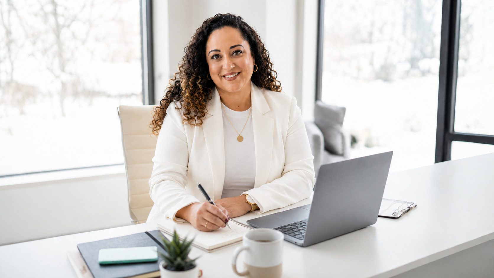 A woman with curly hair, wearing a white blazer and a gold necklace, sitting at a white desk in front of a laptop and a notepad, smiling at the camera in a modern office with large windows.