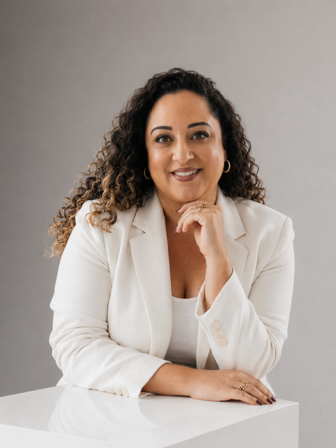 Portrait of a woman with long curly hair, wearing a white blazer and blouse, sitting at a white table with a gray background.