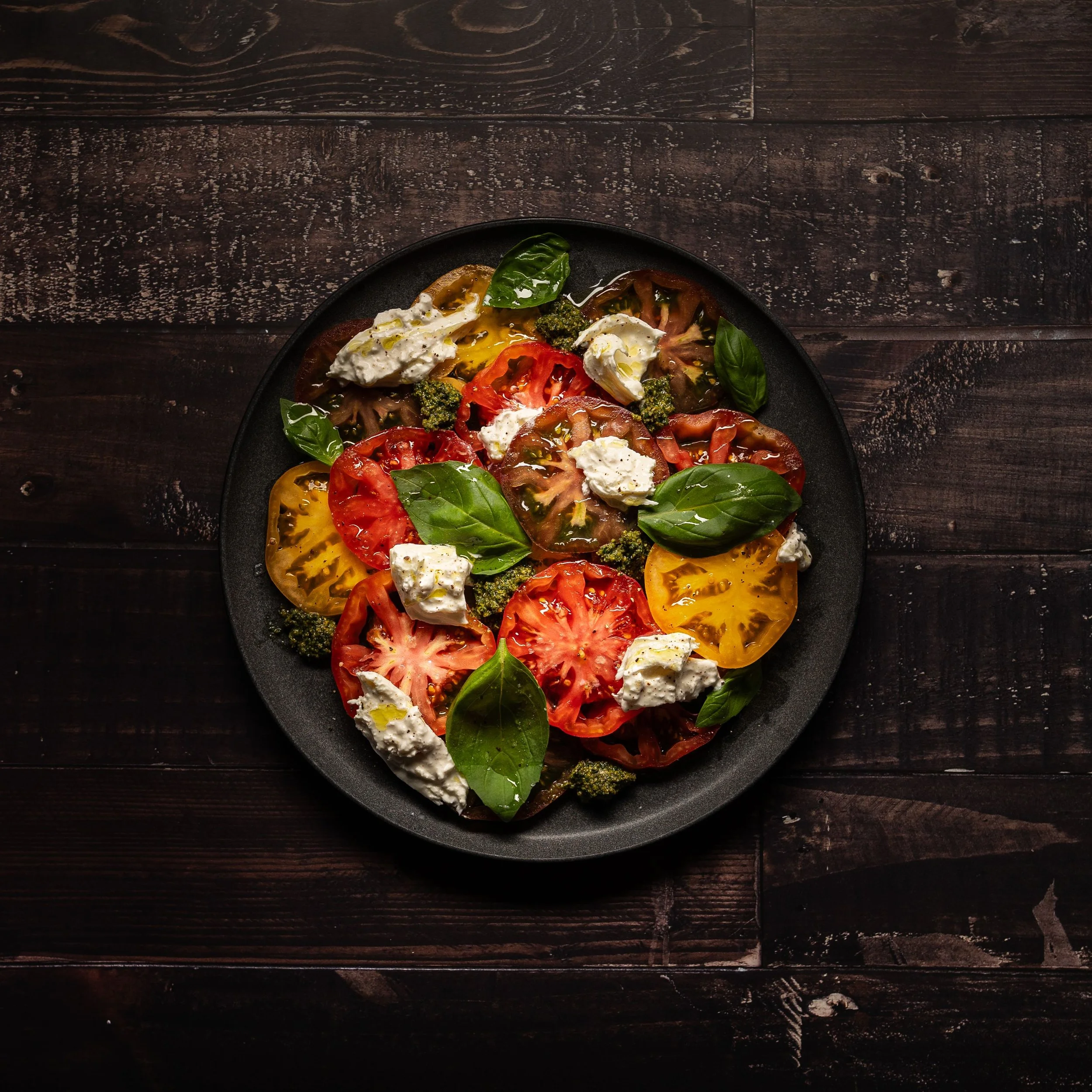 A fresh twist on a Caprese salad, thin cut tomatoes, ricotta cheese, and basil drizzled with fresh herbs and olive oil, well lit against a dark plate letting the food's natural colors pop.